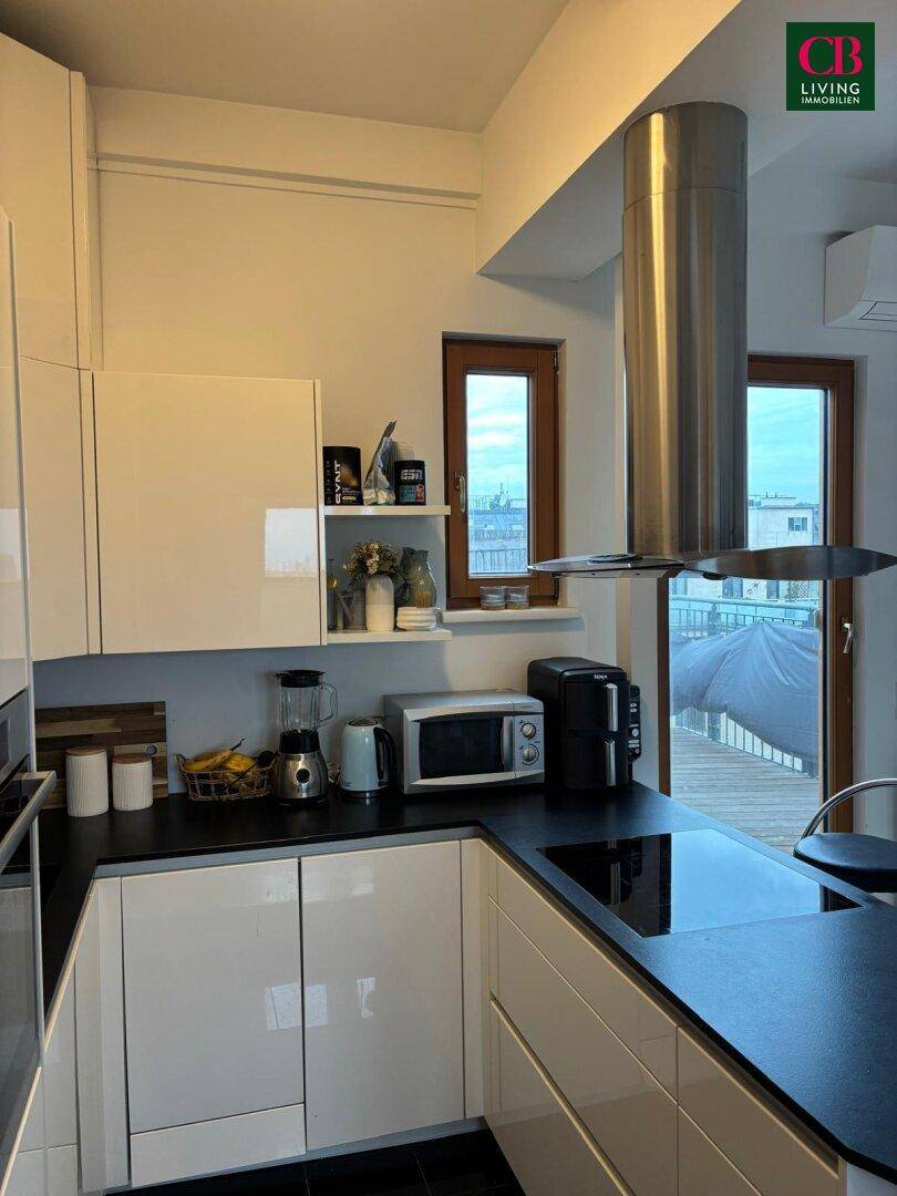 Helle Küche mit weißen Hochglanzschränken, dunkler Arbeitsplatte und Fenstern mit Blick nach draußen.