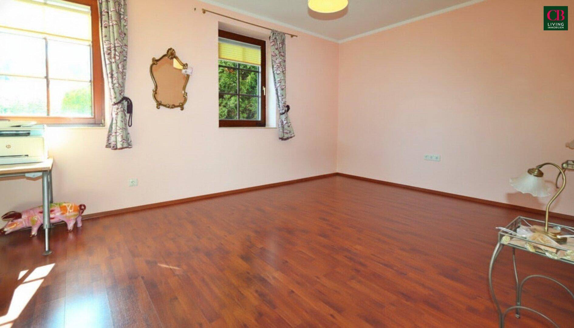 Geräumiges Zimmer mit Holzboden, hellrosa Wänden und zwei Fenstern mit Blick ins Grüne.