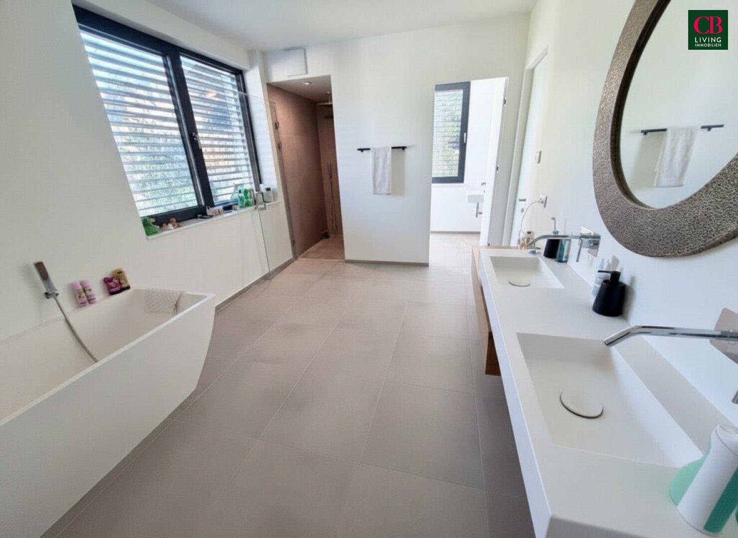 Modernes Badezimmer mit freistehender Badewanne, Doppelwaschbecken und großem Spiegel, hell gefliest.