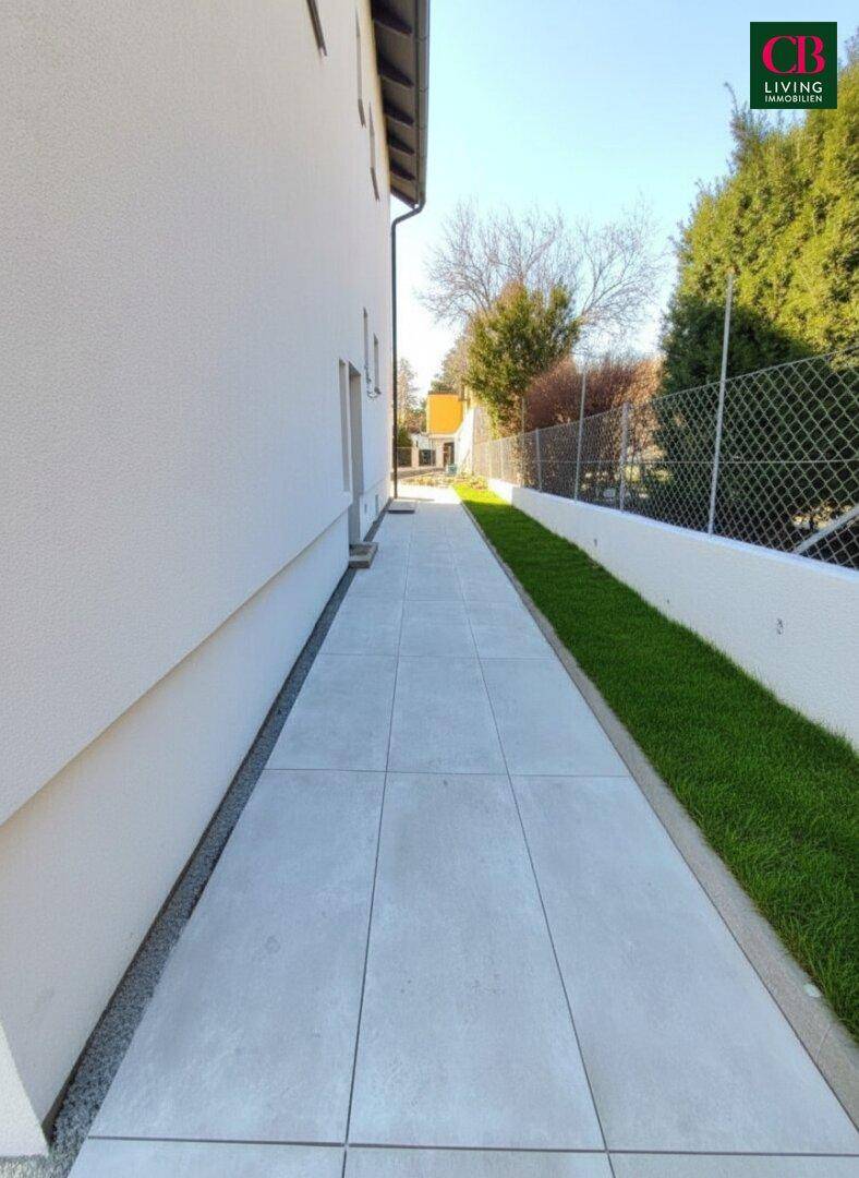 Gepflasterter Seitenweg entlang des Hauses mit einer schmalen Rasenfläche und Zaun.