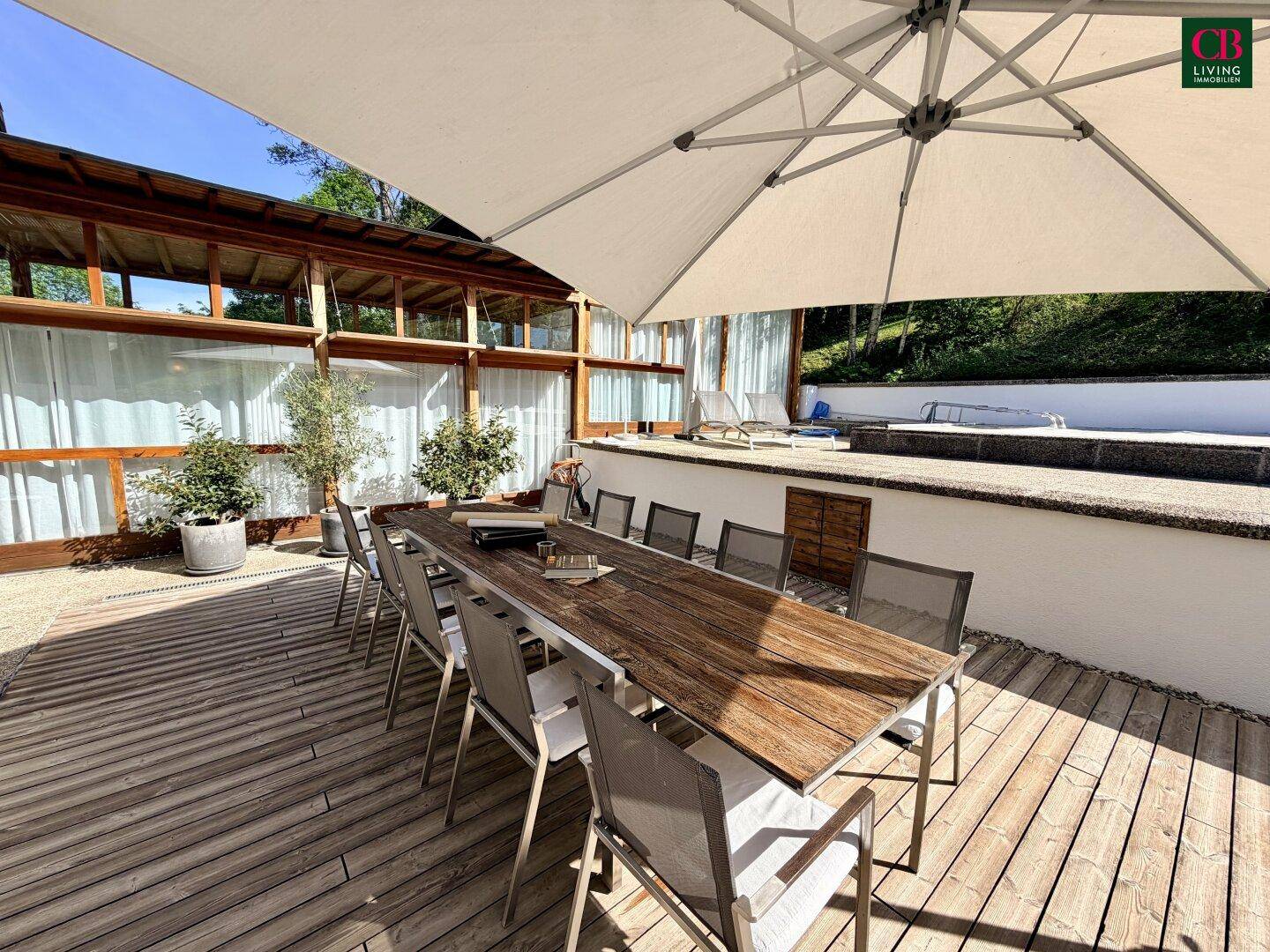 Geräumige Holzterrasse mit großem Esstisch und Stühlen unter einem Sonnenschirm, umgeben von Pflanzen.