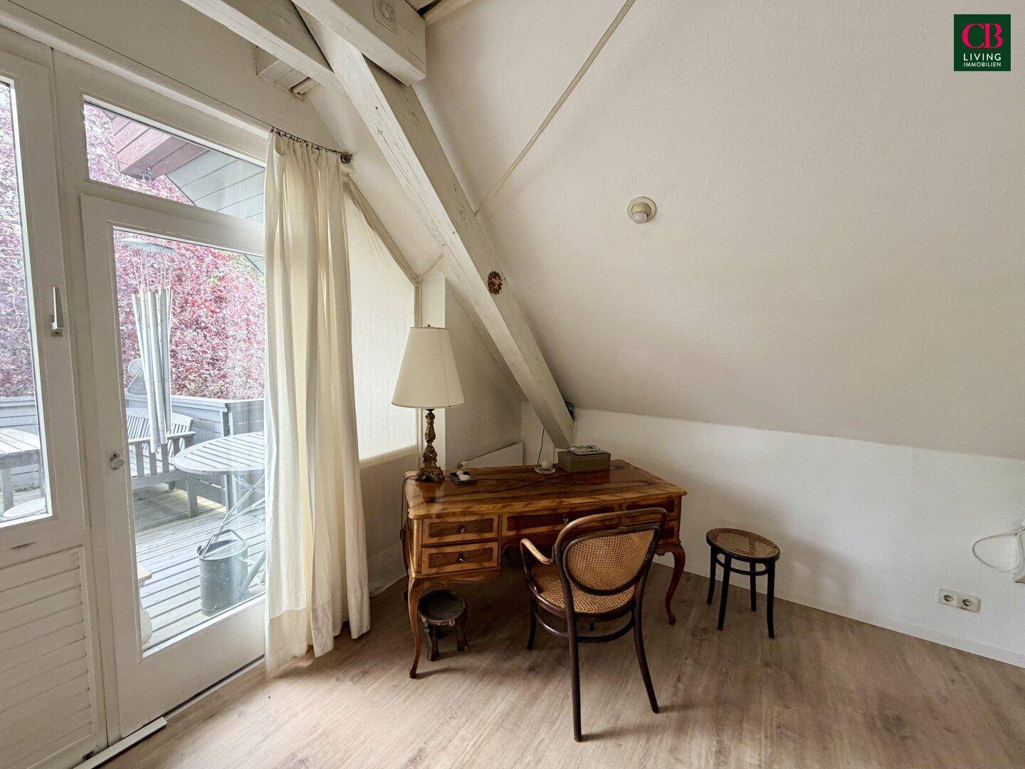 Arbeitsbereich im Dachgeschoss mit Holztisch, Stuhl und Zugang zu einem kleinen Balkon.