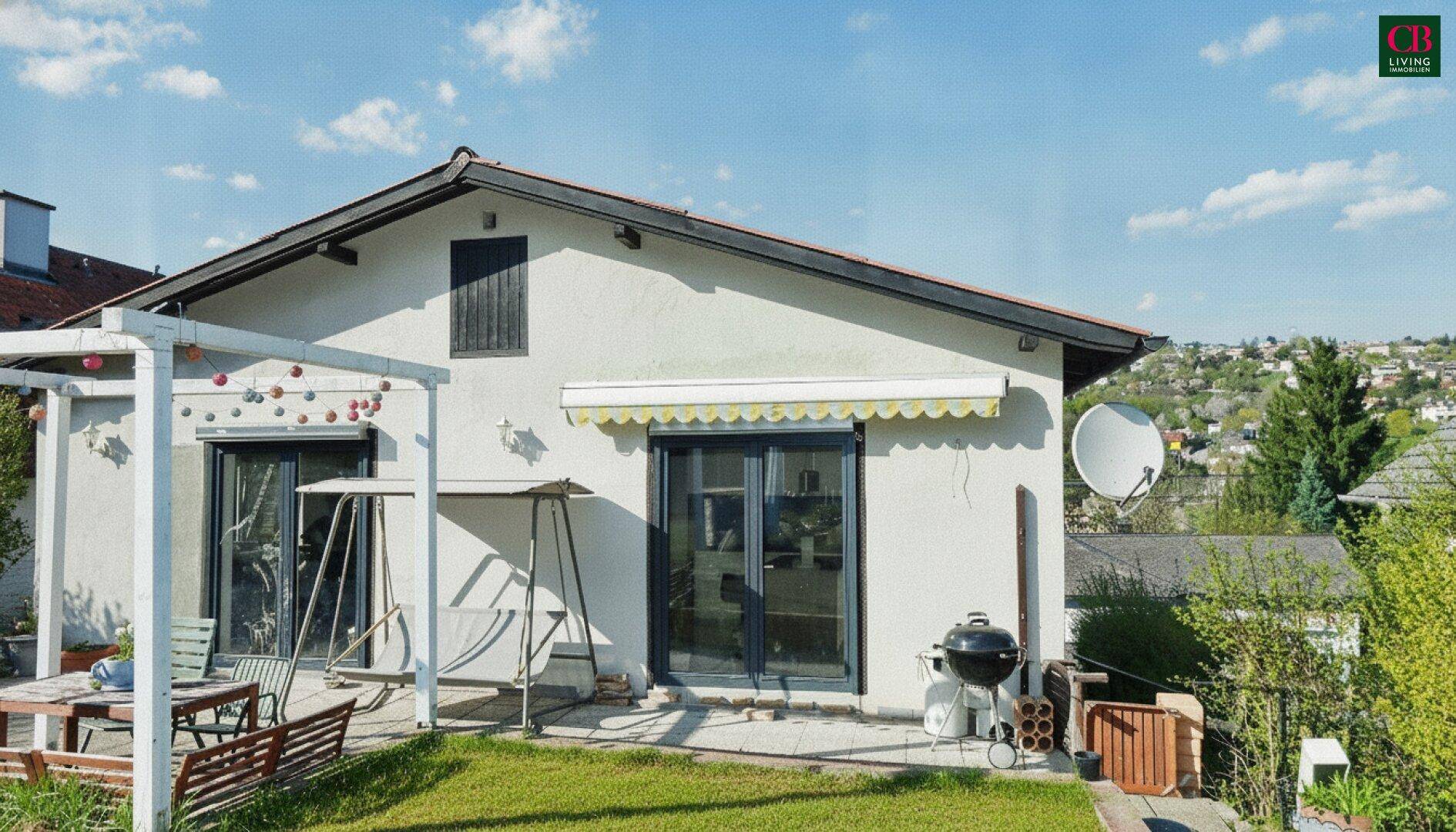 Hausfassade mit Terrasse, Gartenmöbeln, Grill und Pergola unter blauem Himmel.