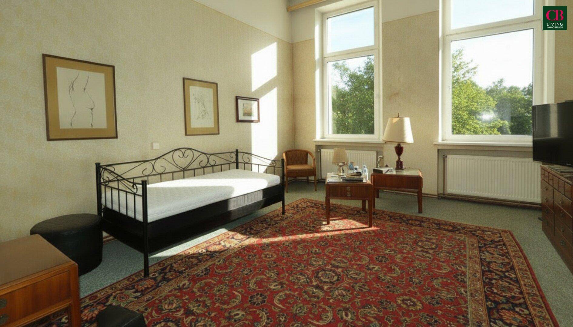 Helles Schlafzimmer mit Tagesbett, Teppich und Blick ins Grüne durch große Fenster.