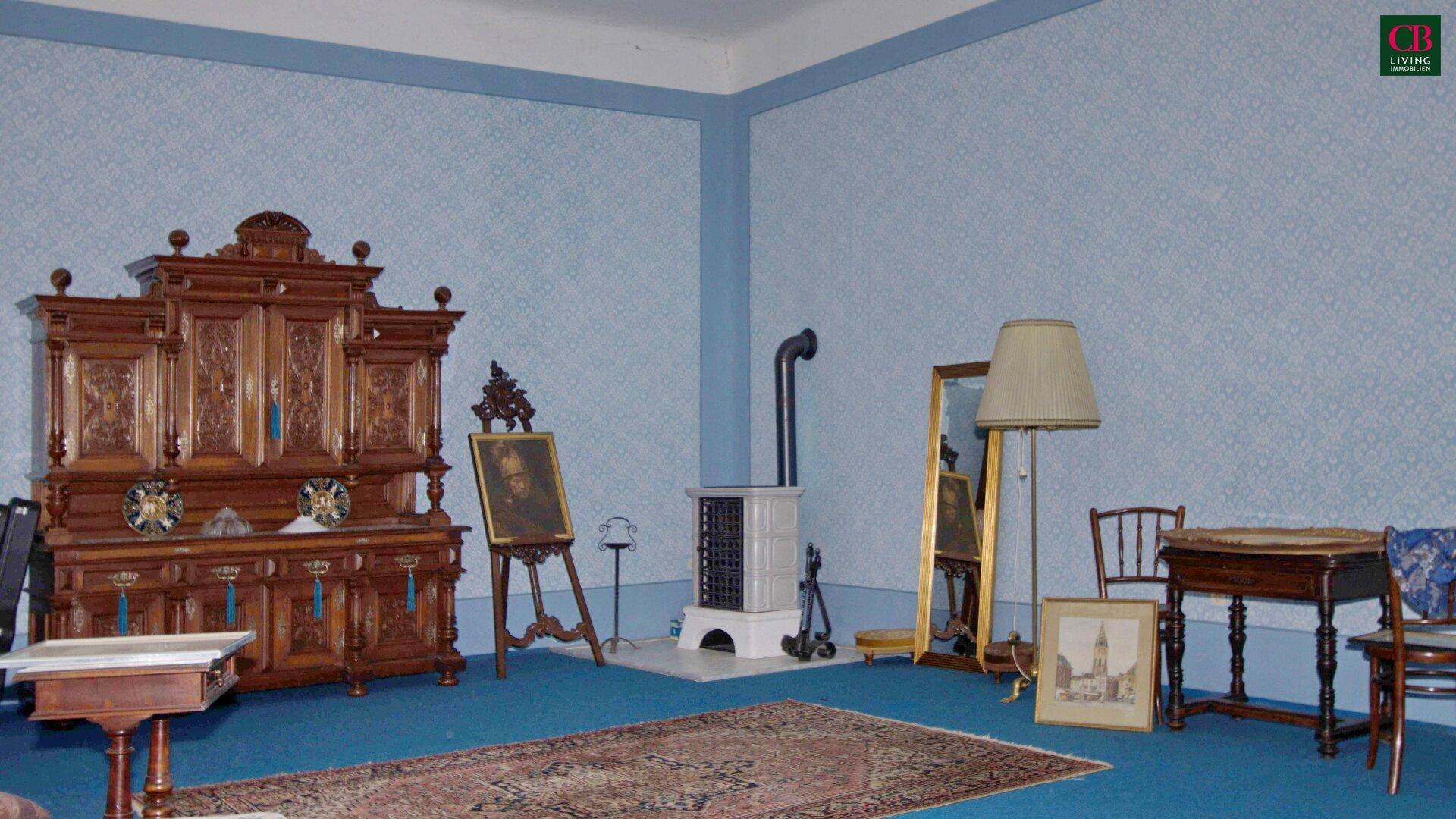 Wohnzimmer mit antiken Möbeln, Kaminofen und blauer Tapete, durchflutet von Tageslicht.