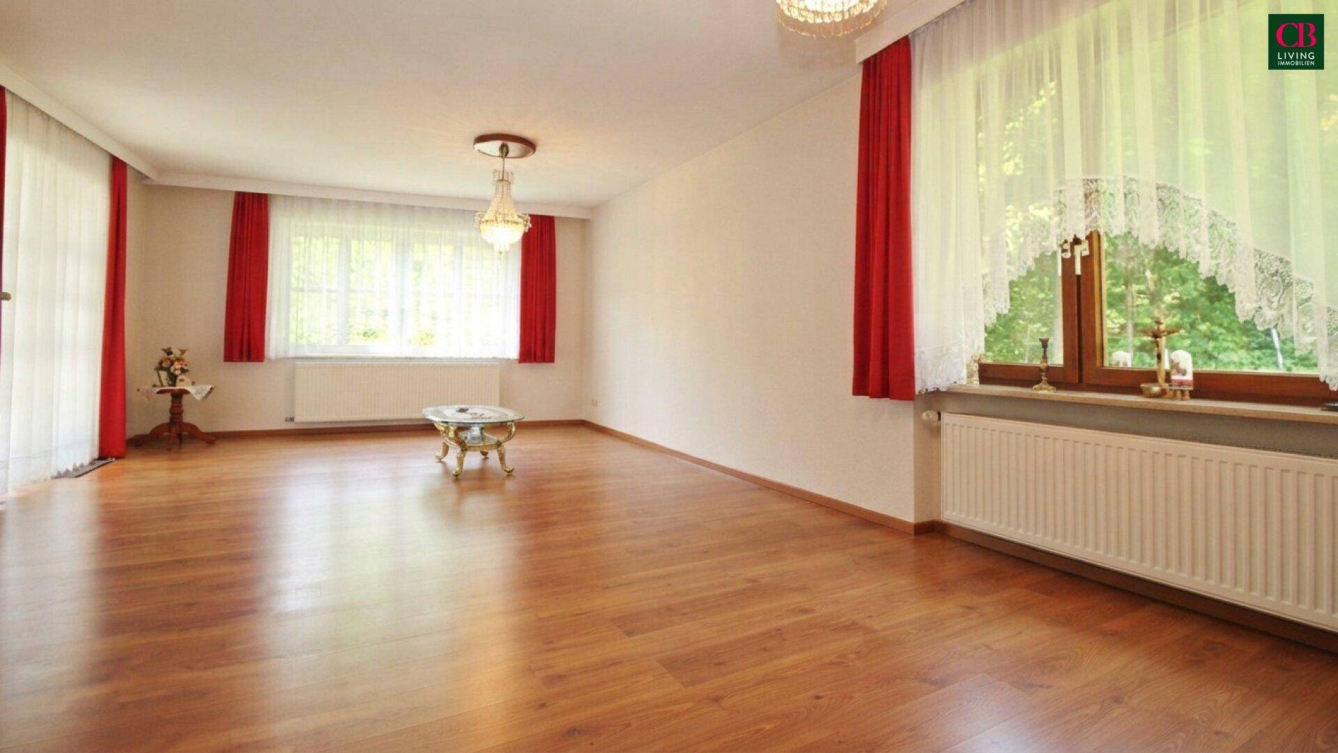 Geräumiges Wohnzimmer mit Holzboden, großen Fenstern und klassischer Deckenleuchte.
