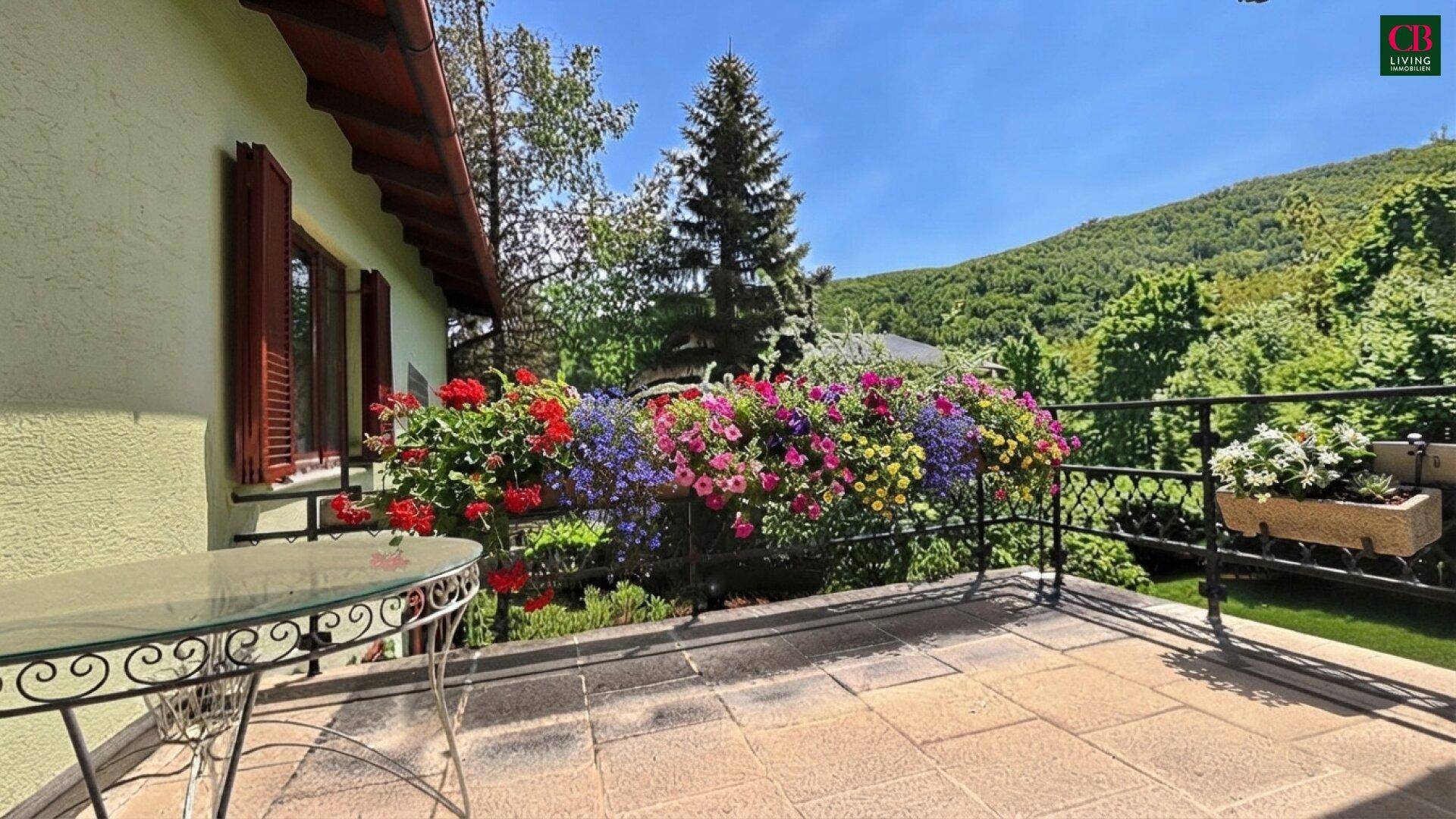 Große gepflasterte Terrasse mit Tisch und bunten Blumen vor grüner Hügellandschaft.
