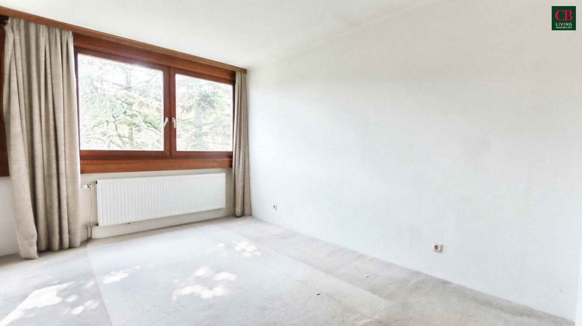 Leeres Zimmer mit hellem Teppichboden, weißen Wänden und Fenster mit Blick ins Grüne.