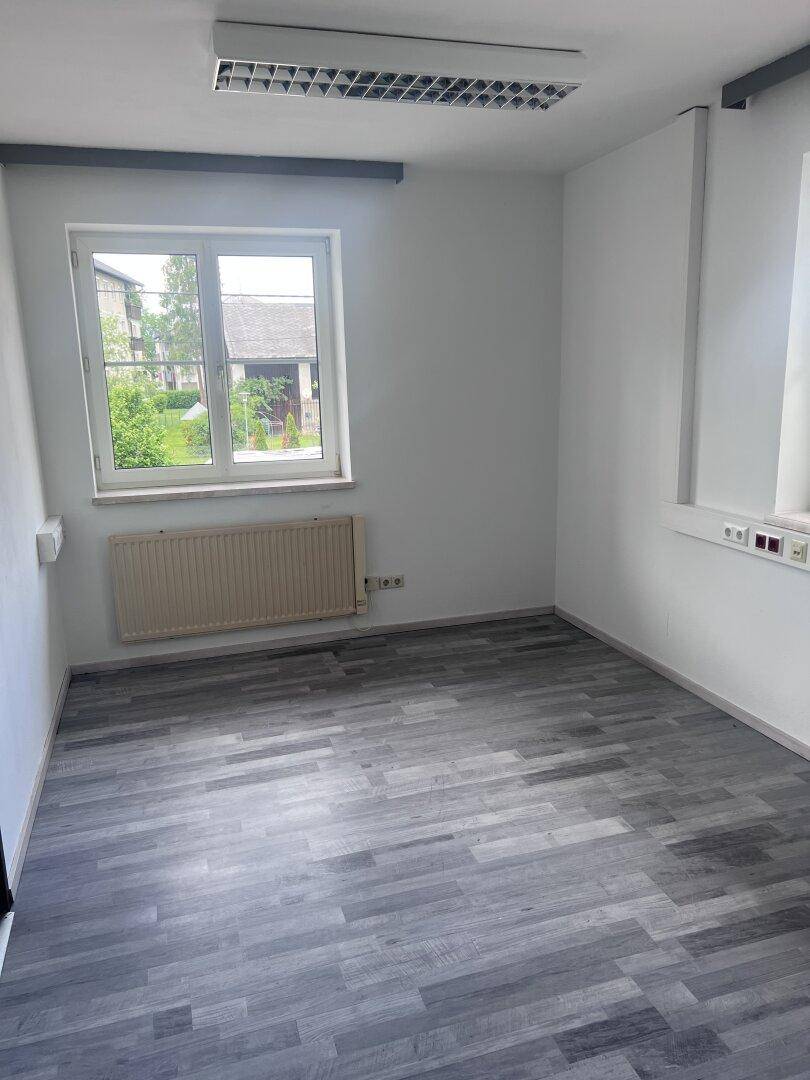 Heller Büroraum mit grauem Laminatboden, einem großen Fenster und einem Heizkörper darunter.