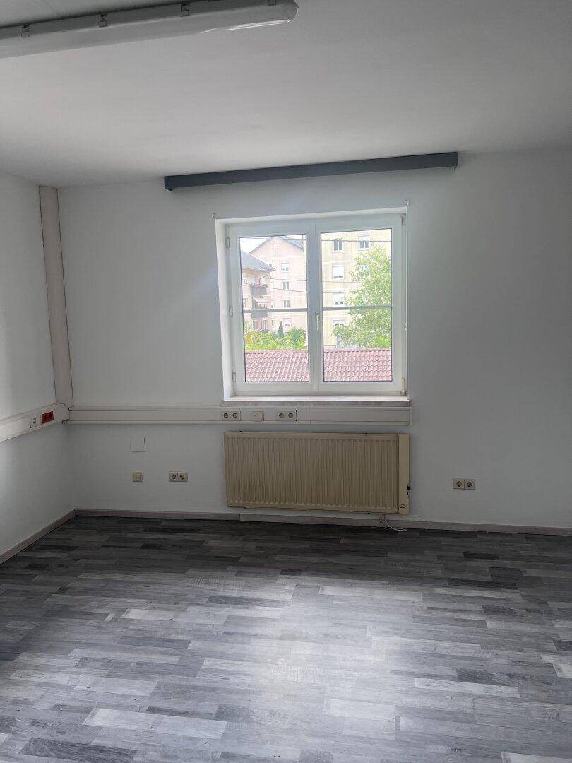 Leerer Büroraum mit grauem Laminatboden, einem Fenster und einem Heizkörper an der Wand.