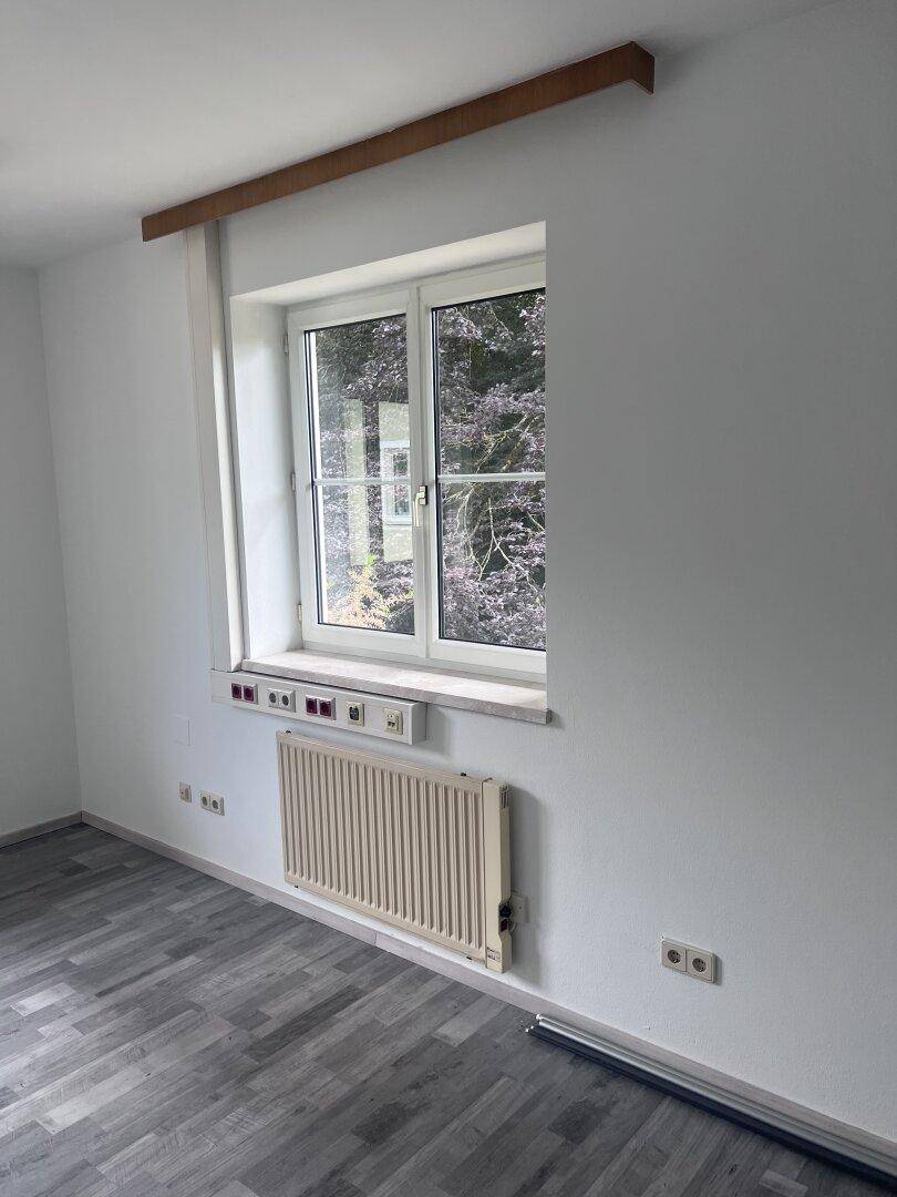 Büroraum mit grauem Laminatboden, einem Fenster und einem Heizkörper unter der Fensterbank.