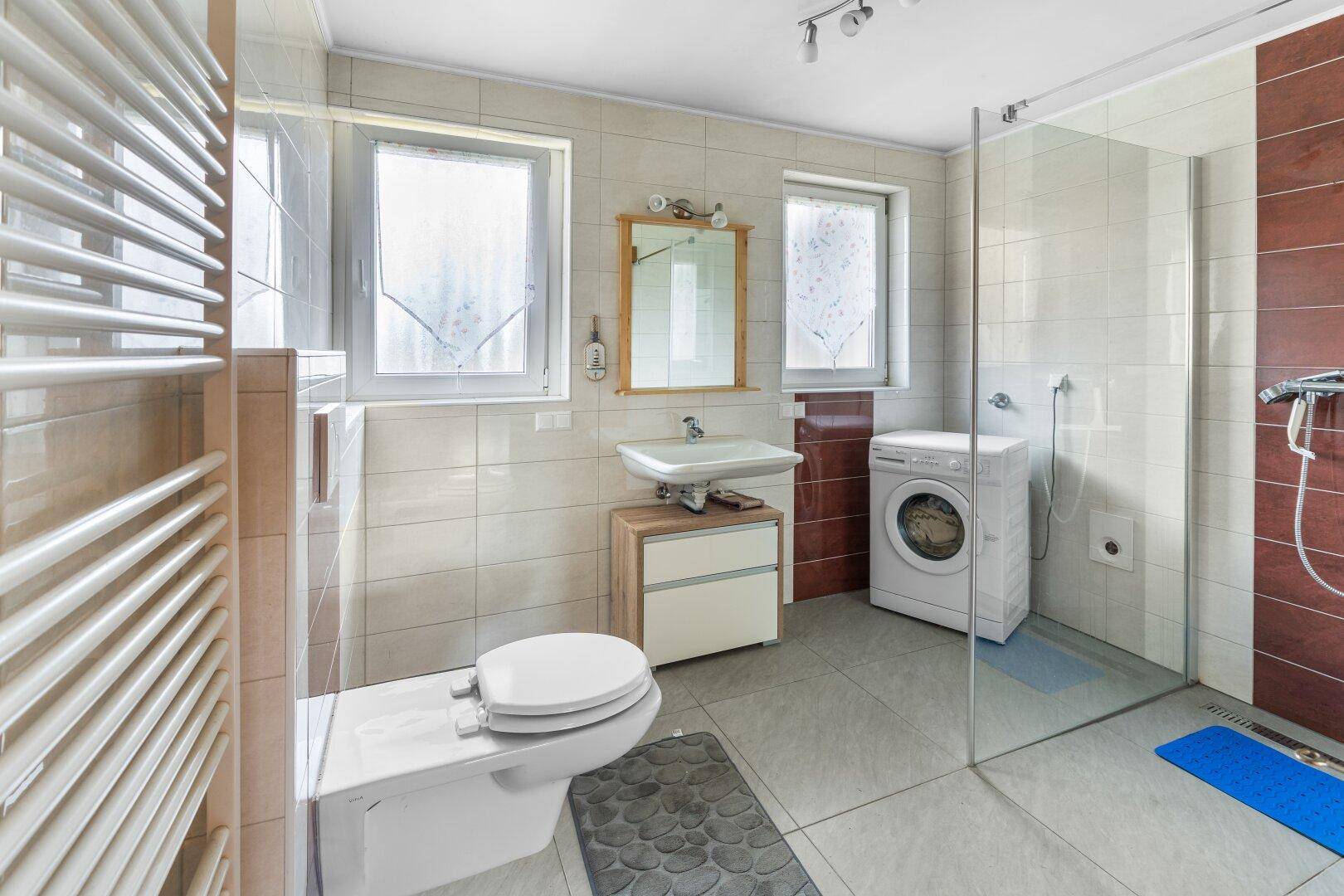 Helles Badezimmer mit Duschkabine, Toilette, Waschbecken und Waschmaschine, gefliest.