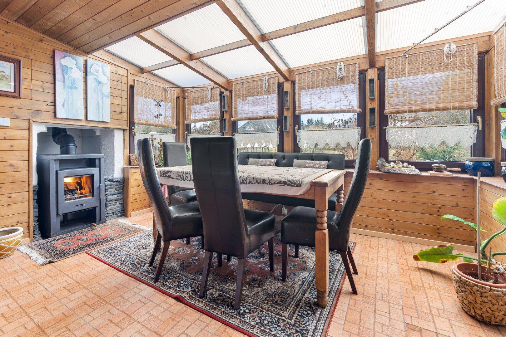 Gemütlicher Wintergarten mit Holzofen, Esstisch und viel Tageslicht.