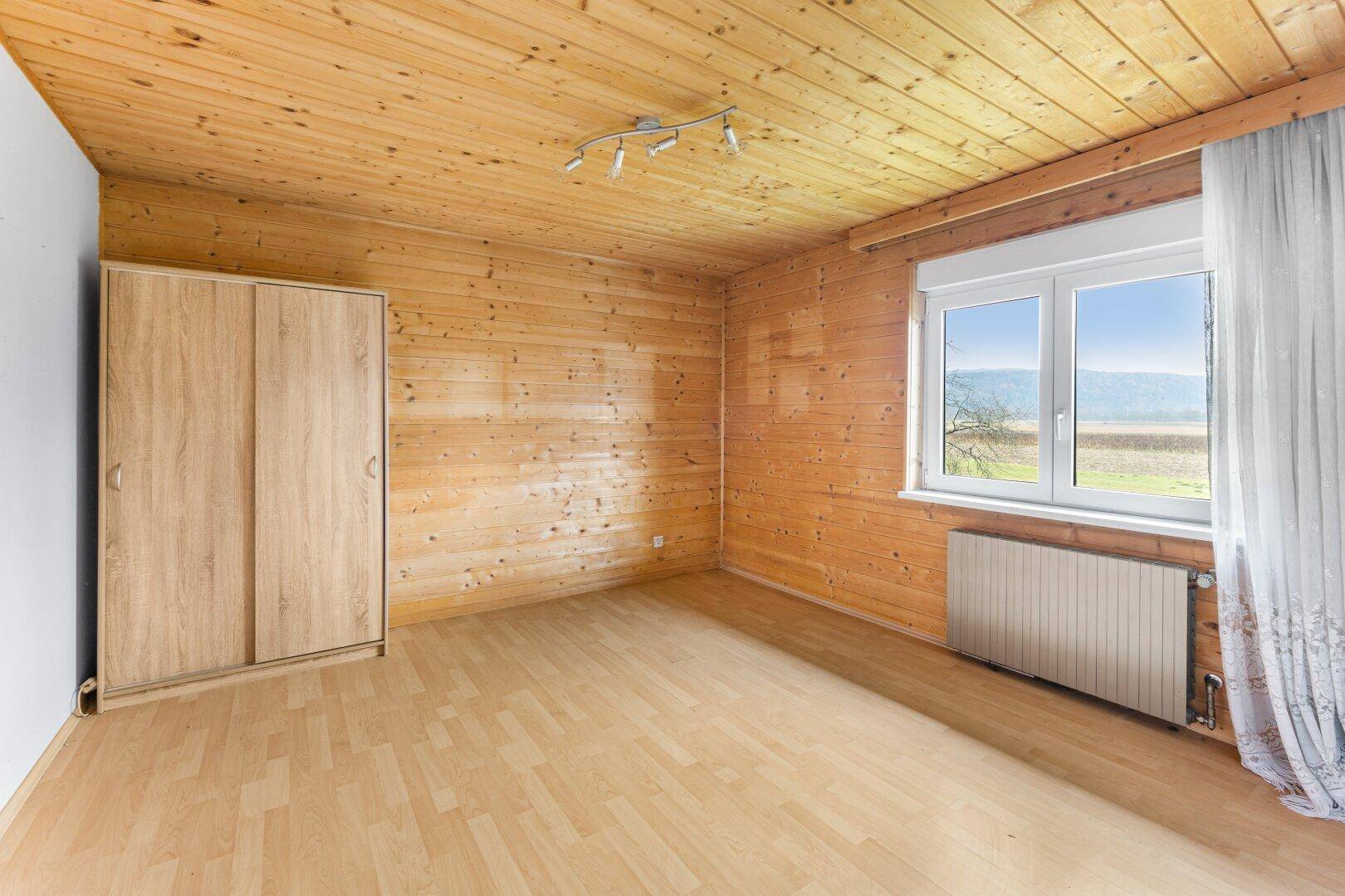 Schlafzimmer mit Laminatboden, Holzvertäfelung und Fenster mit Blick ins Grüne.