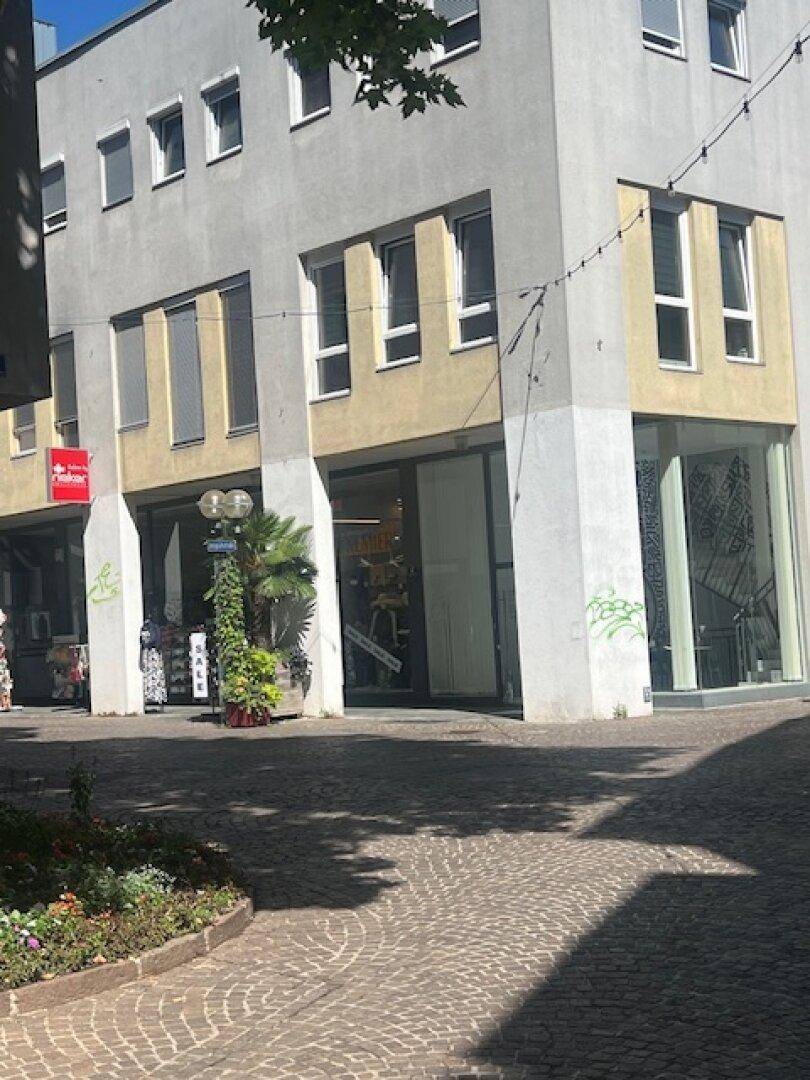 Mehrstöckiges Gebäude mit Geschäften im Erdgeschoss und Wohnungen darüber, Fassade in Grau und Beige mit vielen Fenstern und Rollos.