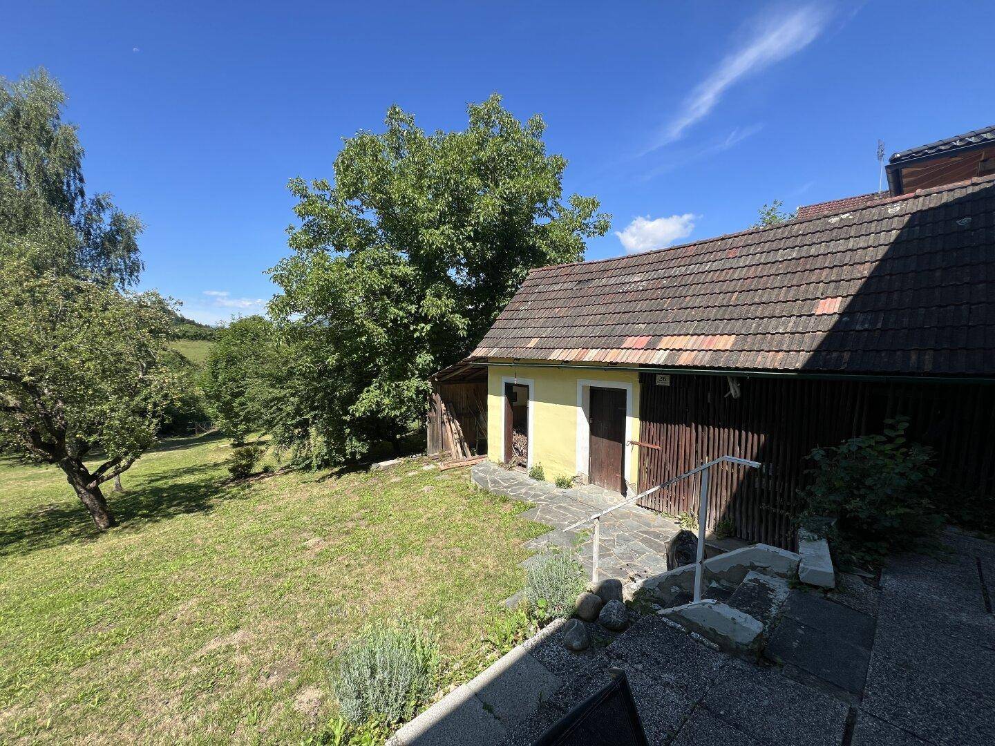 Kleines gelbes Nebengebäude mit braunem Dach und Steintreppe in ländlicher Umgebung.