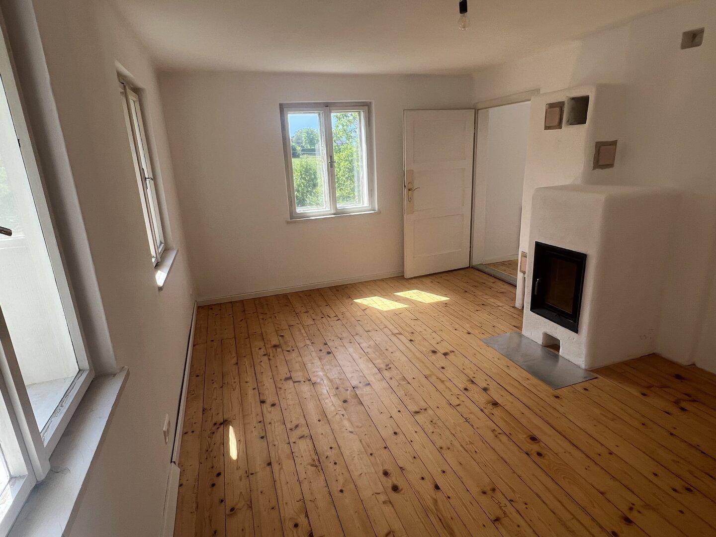 Heller Wohnraum mit Holzfußboden, zwei Fenstern und einem modernen Kaminofen.