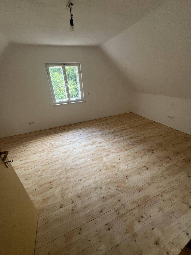 Helles Dachzimmer mit schrägen Wänden, Holzfußboden und einem Fenster mit Blick ins Grüne.