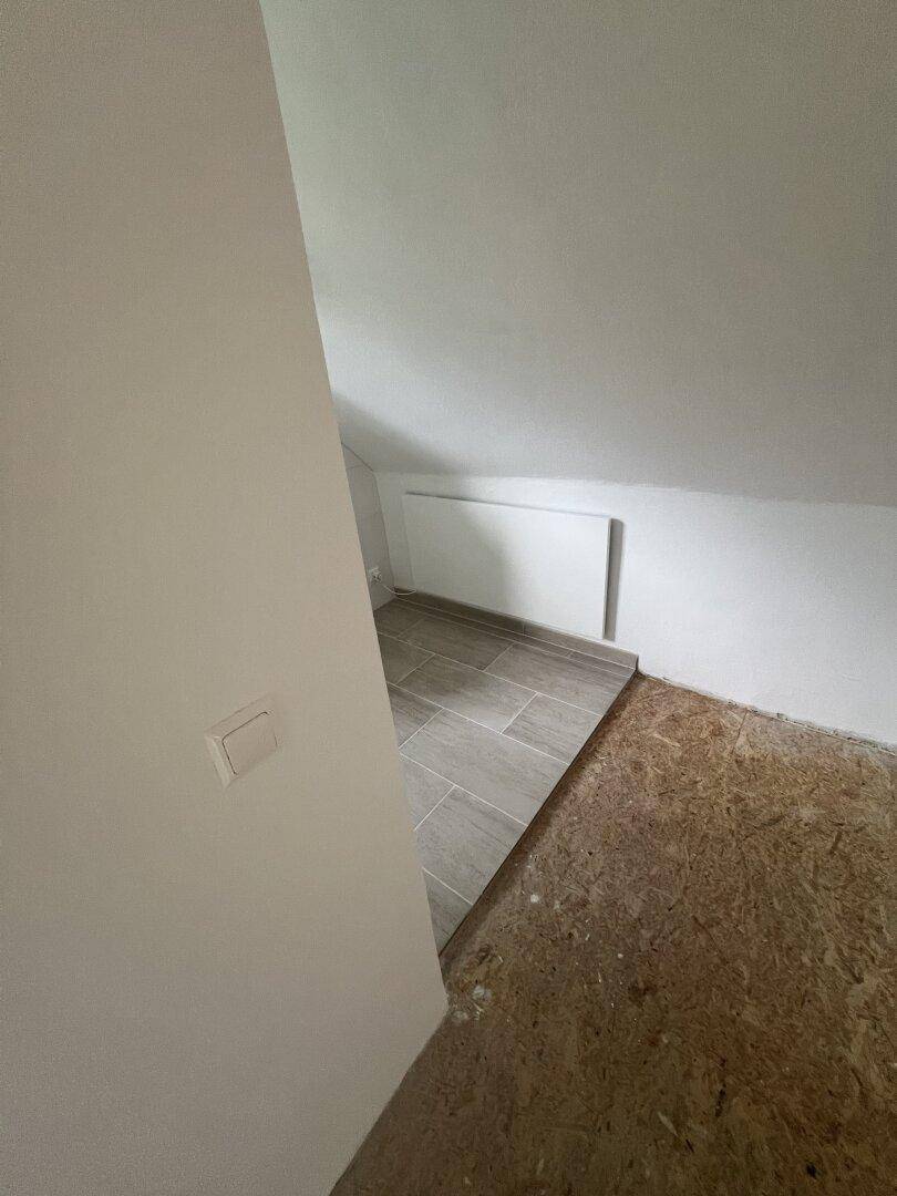Ecke eines Dachzimmers mit schräger Decke, Heizkörper und gemischtem Bodenbelag.