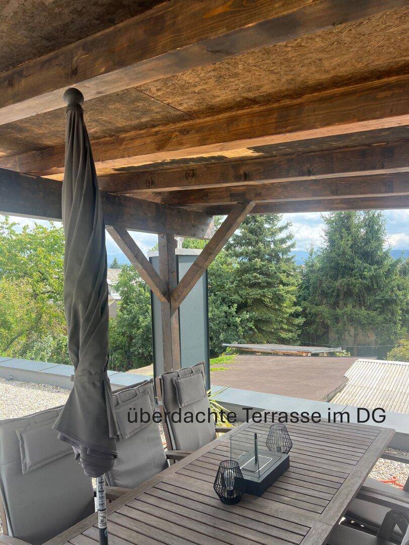 Überdachte Terrasse im Dachgeschoss mit Holzbalken und Gartenmöbeln, umgeben von Bäumen.