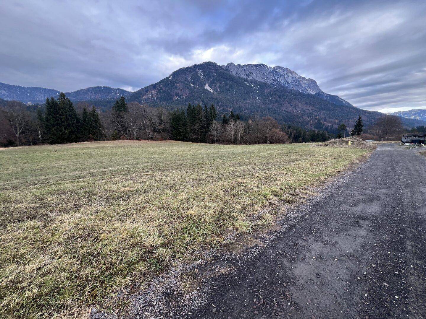 Weite Wiesenfläche mit majestätischem Bergblick und einem Schotterweg am Rand.
