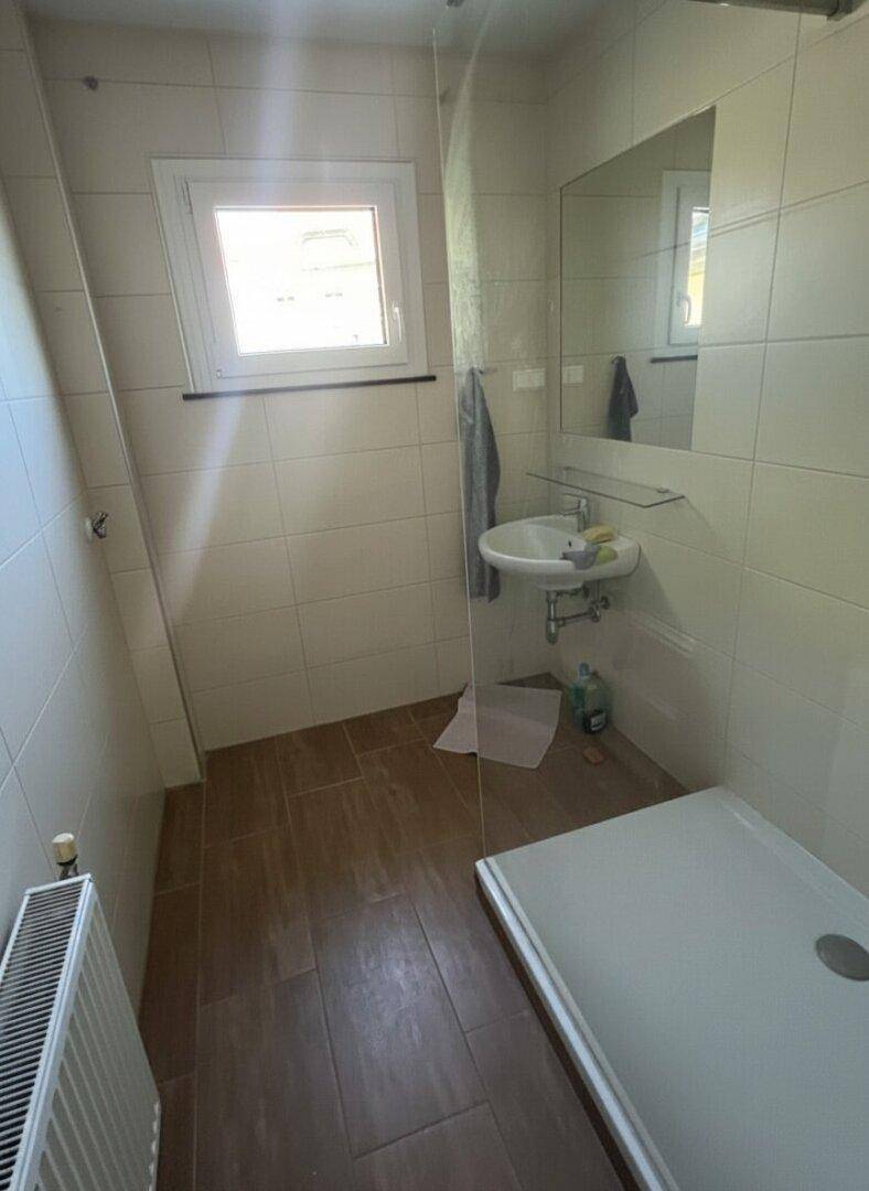 Badezimmer mit Dusche, Waschbecken, Fenster und Heizkörper, hell gefliest.