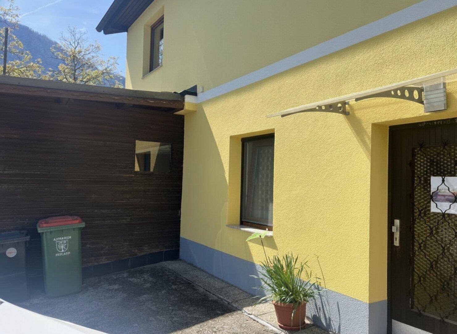 Seitenansicht des gelben Hauses mit Eingangsbereich und Mülltonnen.