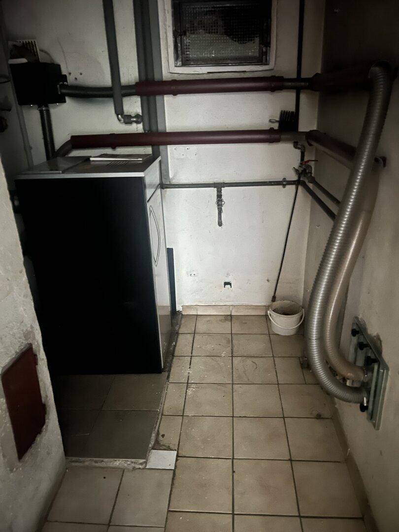 Funktionaler Waschraum im Keller mit Waschmaschine, Fliesenboden und sichtbaren Installationen.