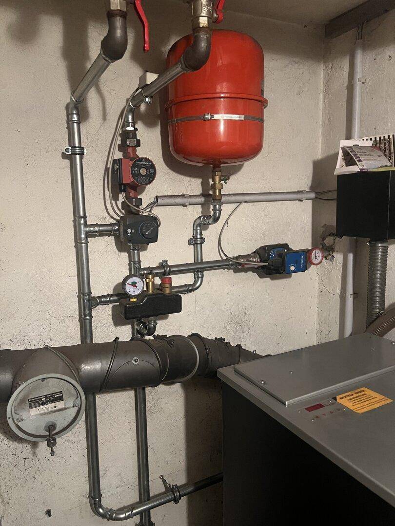 Heizungsraum mit komplexem Rohrsystem und einem roten Warmwasserspeicher im Kellerbereich.