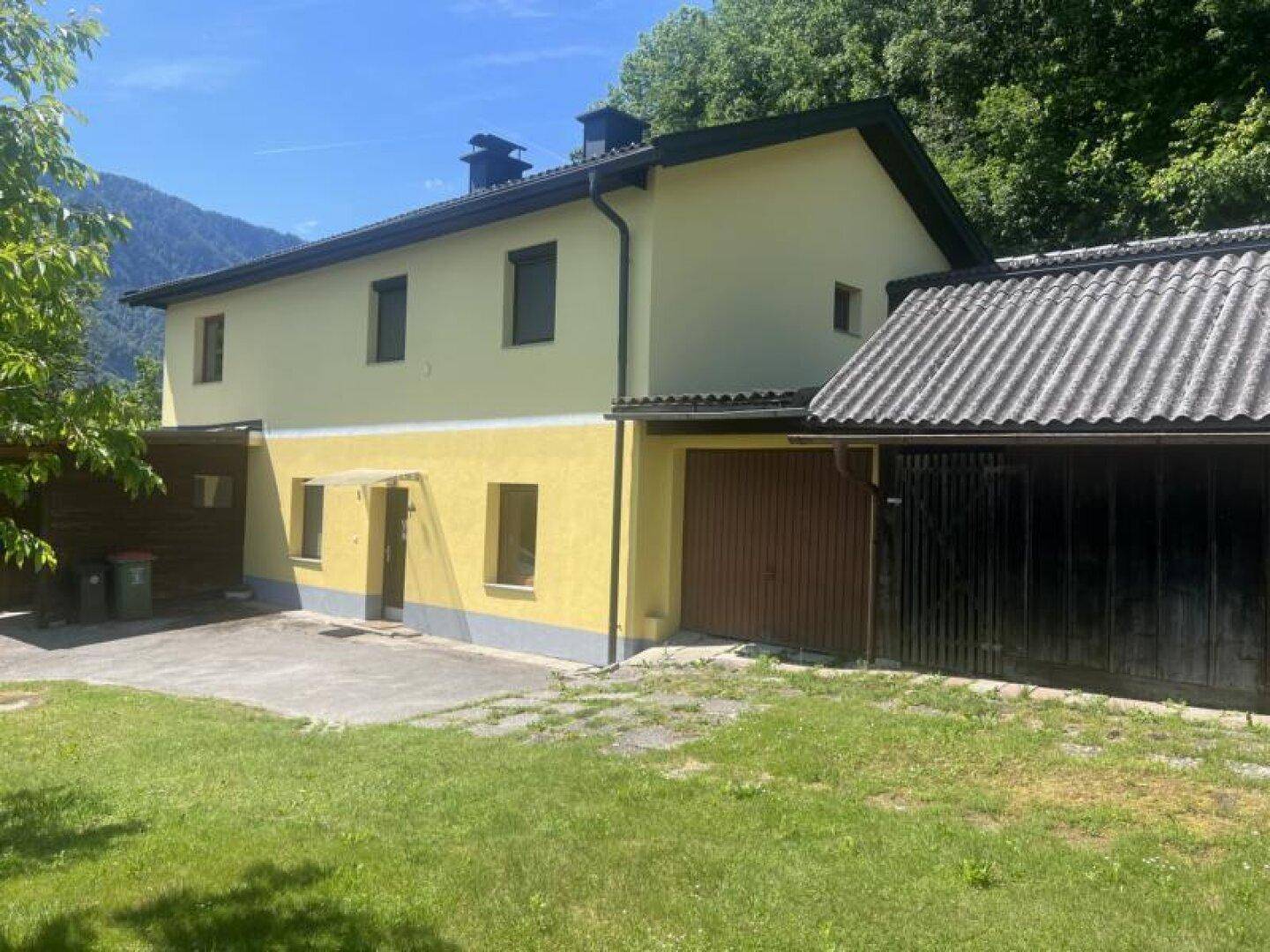 Gelbes Haus mit dunklem Satteldach und angrenzender Garage, umgeben von viel Grün und Bergen im Hintergrund.
