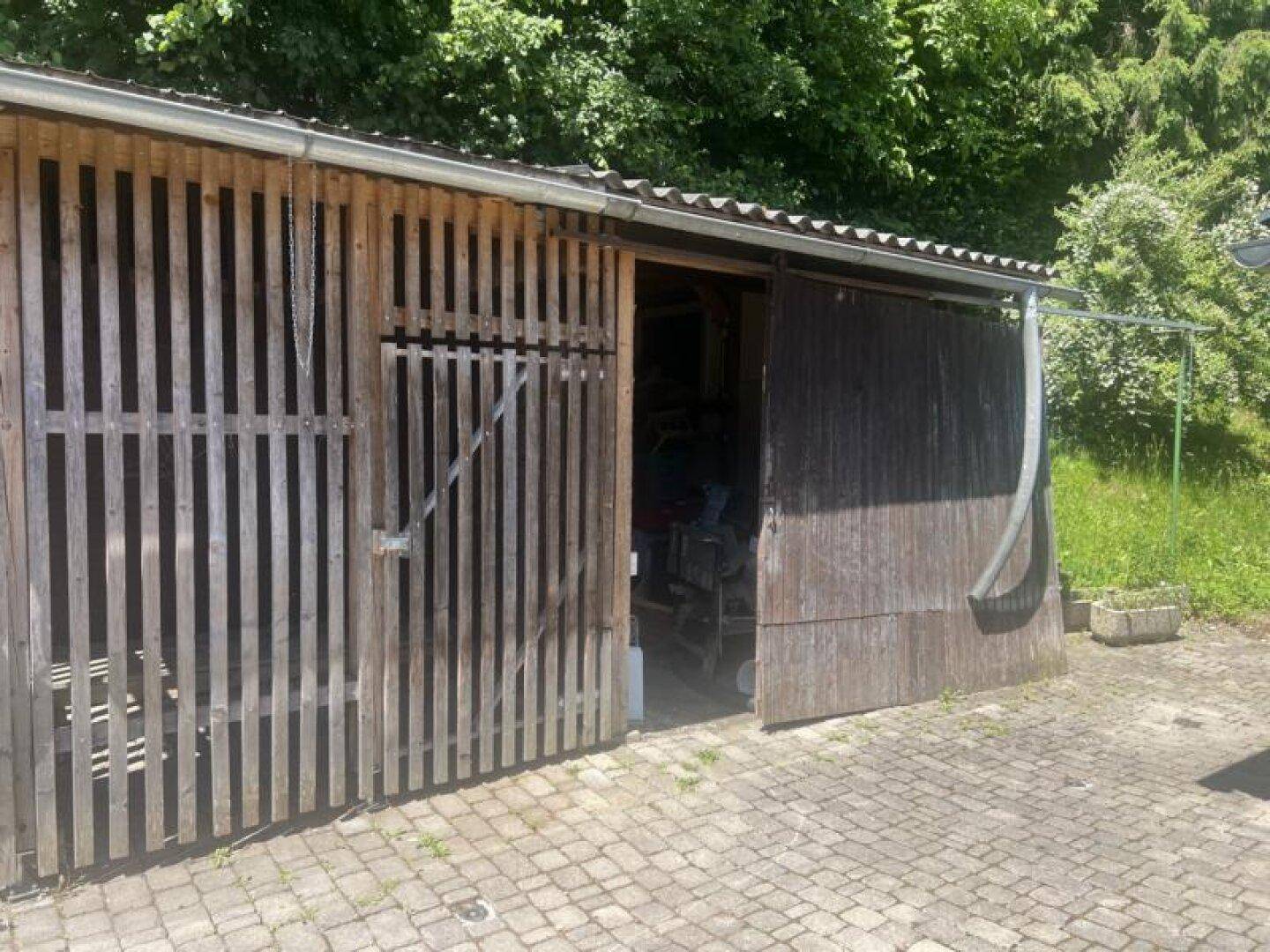 Rustikaler Holzschuppen mit Wellblechdach und gepflastertem Vorplatz, umgeben von Bäumen und Grün.