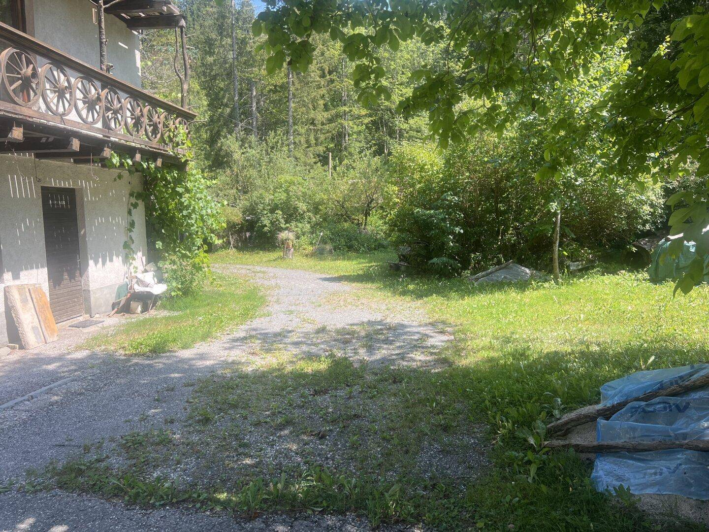Kiesweg und Rasenfläche vor einem Gebäude mit traditionellem Holzbalkon, umgeben von üppigem Grün.