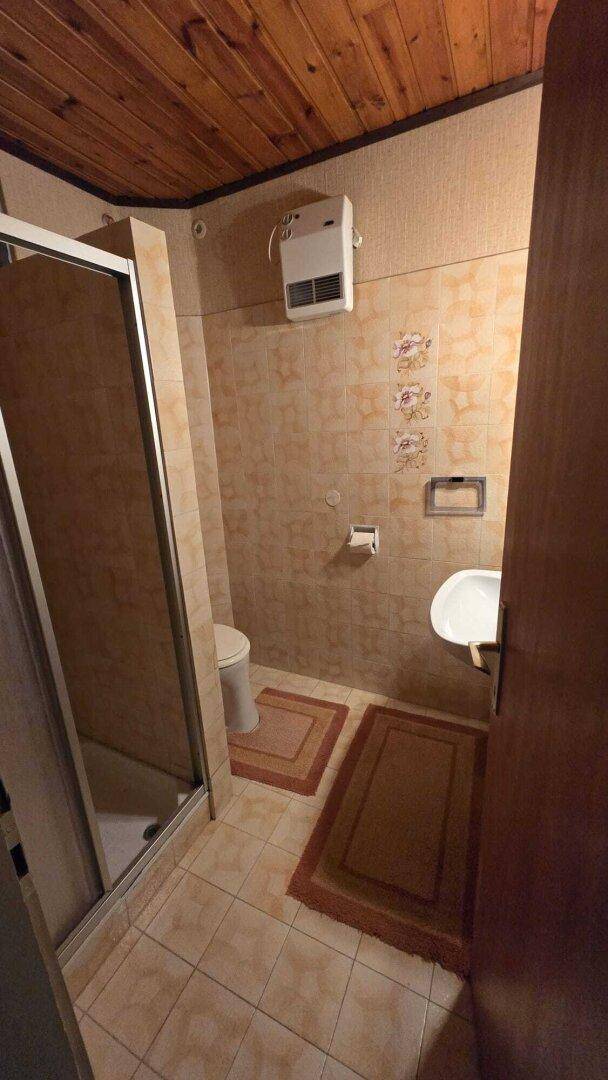 Kleines Badezimmer mit beigefarbenen Fliesen, Duschkabine, WC und einer Holzpaneeldecke.