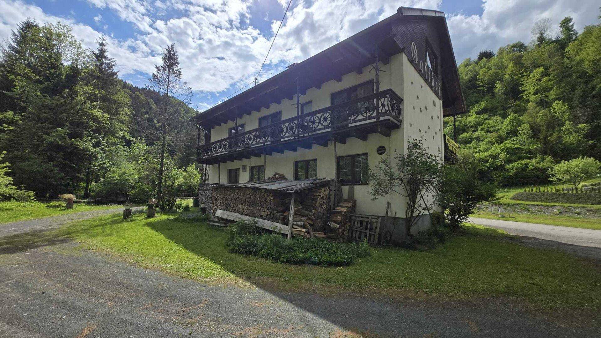 Traditionelles Alpenhaus mit Balkonen und Holzstapeln, umgeben von grüner Natur und Bergen.