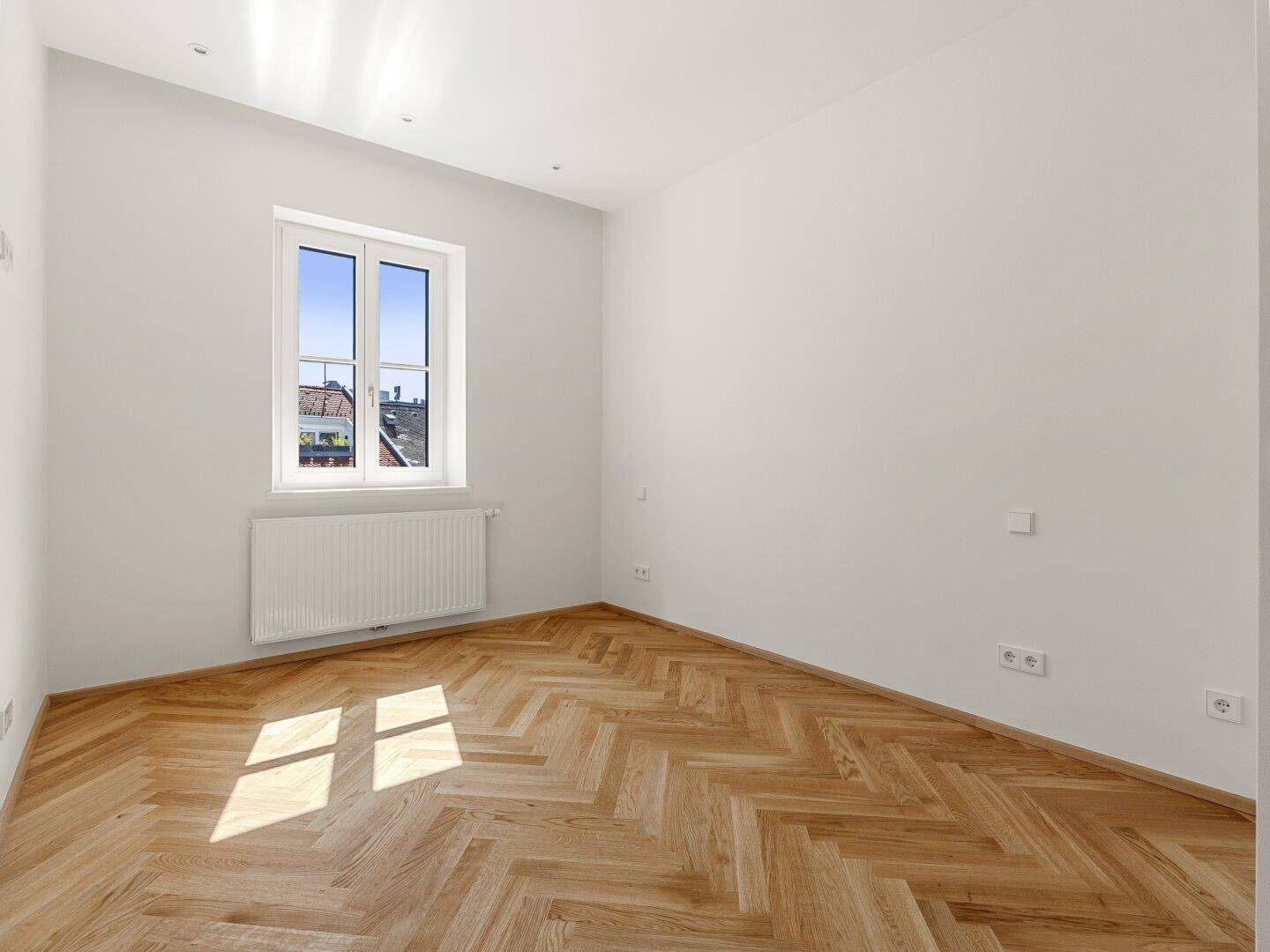 Helles Zimmer mit Fischgrätparkett und einem Fenster, das einen Blick nach draußen bietet.