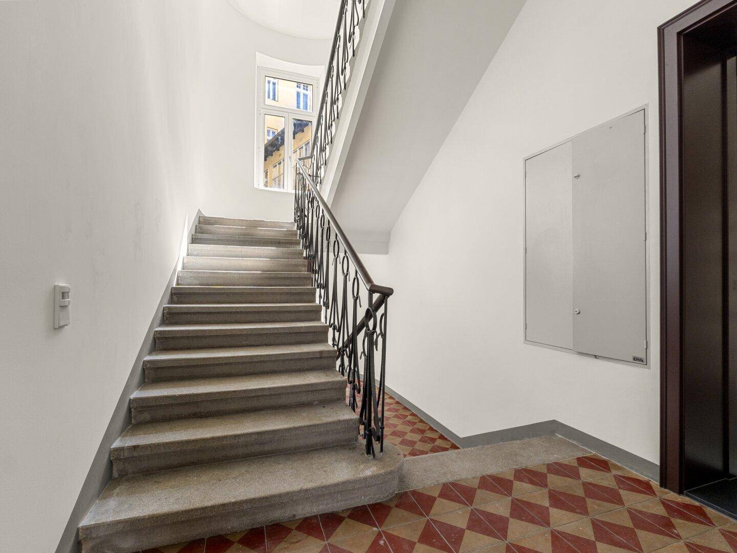 Historisches Treppenhaus mit schmiedeeisernem Geländer und Zugang zu einem modernen Aufzug.