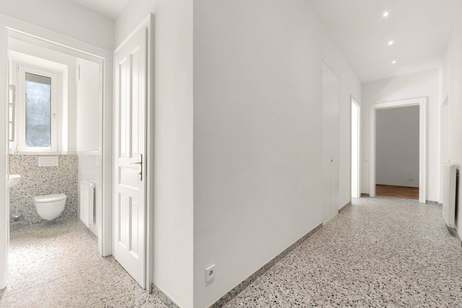 Langer Flur mit Terrazzoboden und Zugang zu einem Badezimmer mit Fenster und wandhängendem WC.