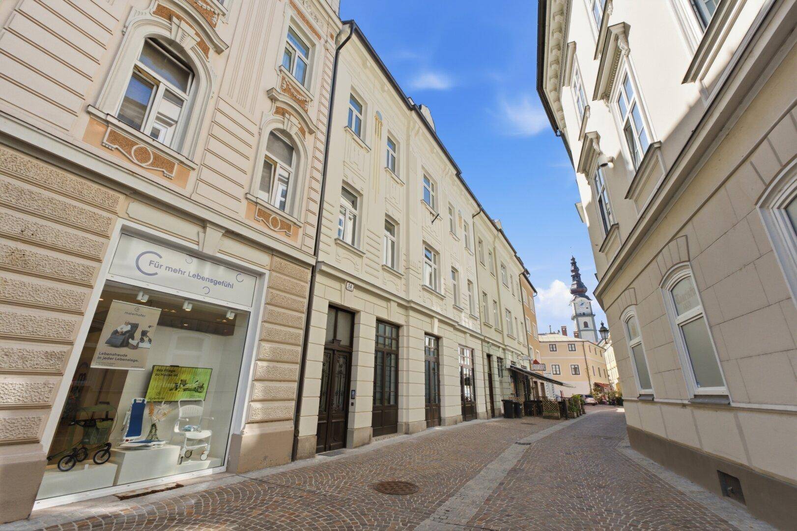 Historische Straße mit Gebäuden und einem markanten Kirchturm im Hintergrund unter blauem Himmel.