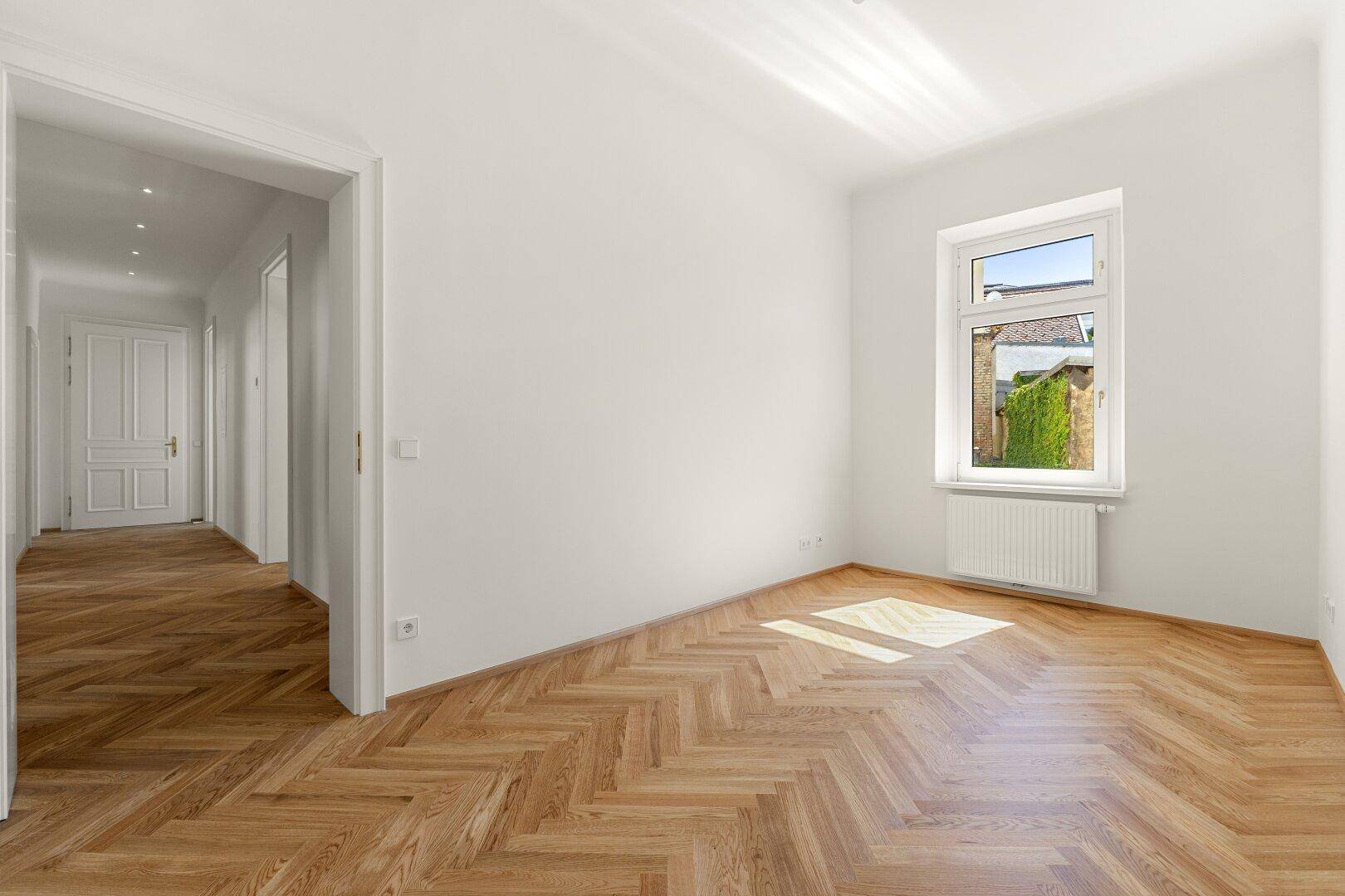 Helles Schlafzimmer mit Fischgrätparkett, weiß gestrichenen Wänden und einem beruhigenden Blick ins Grüne.