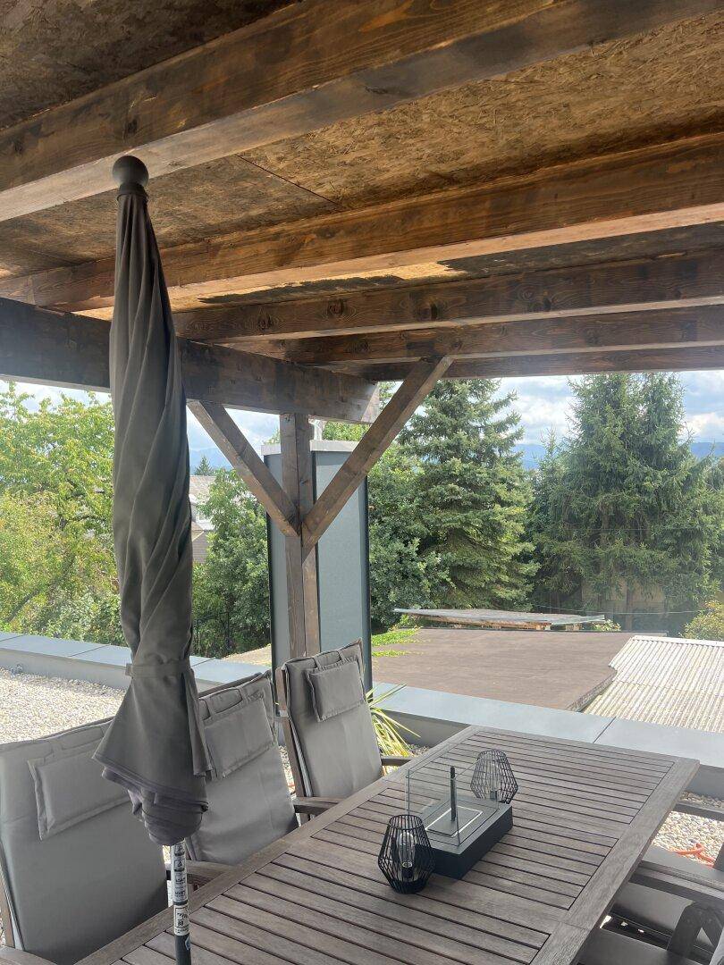 Überdachte Terrasse mit Holztisch und Stühlen, bietet einen weiten Blick ins Grüne.