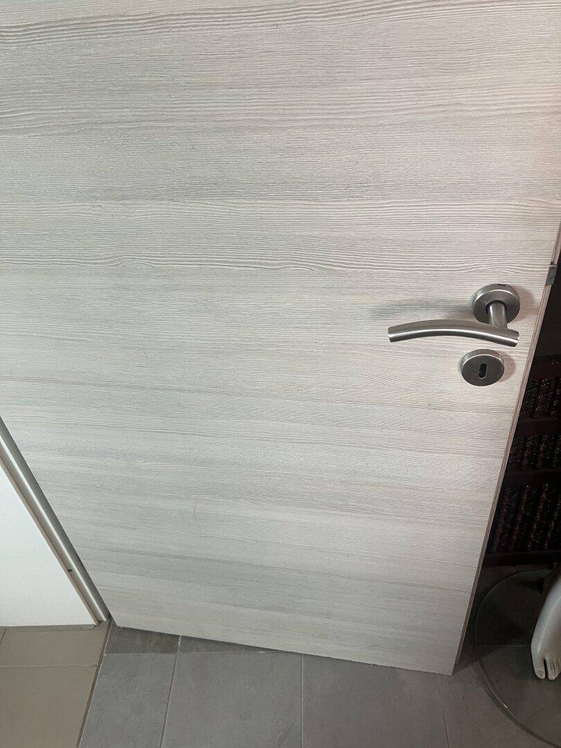 Helle Holztür mit moderner Metallklinke in einem Innenraum mit Fliesenboden.