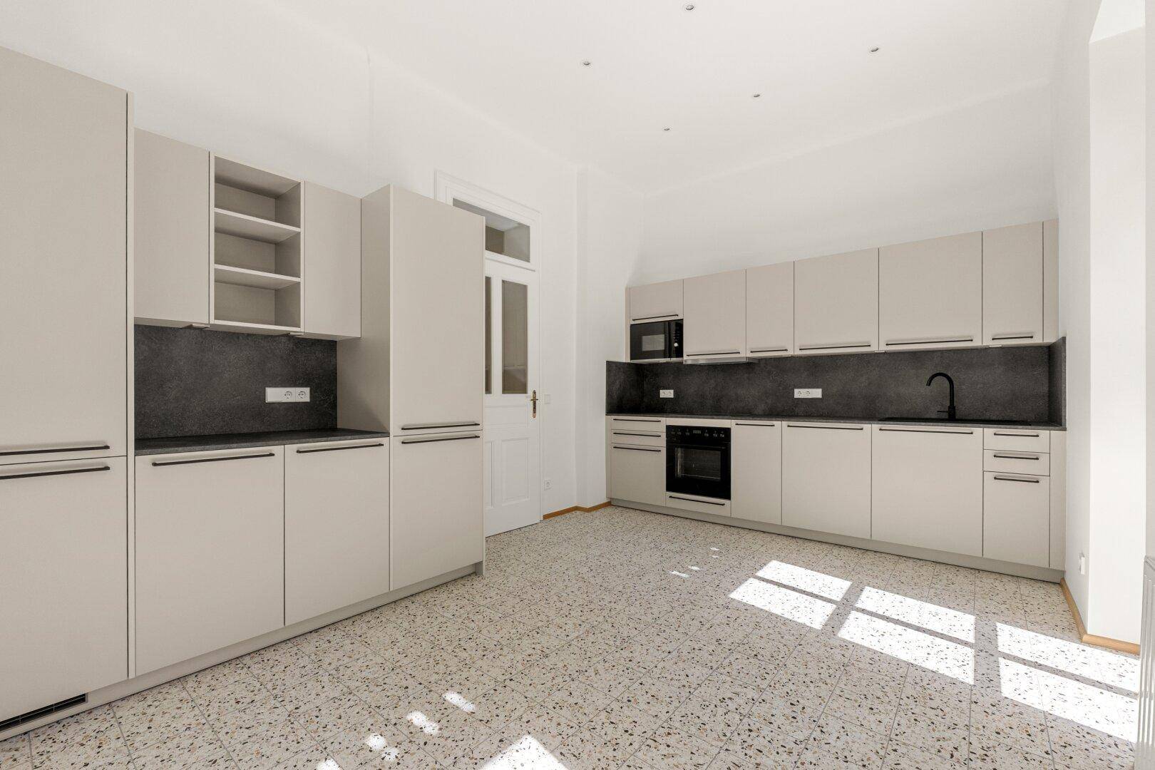 Geräumige, leere Küche mit hellbeigen Schränken, dunkler Arbeitsplatte und Terrazzoboden.