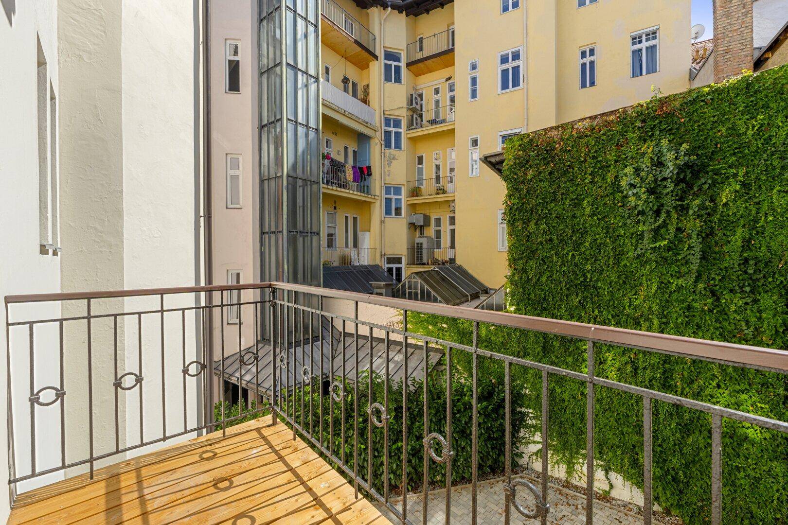 Balkon mit Holzbelag und Metallgeländer, bietet Blick auf den begrünten Innenhof und einen Aufzug.