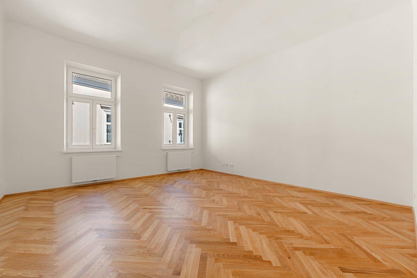 Helles, leeres Zimmer mit Fischgrätparkettboden und zwei großen Fenstern mit Rollläden.