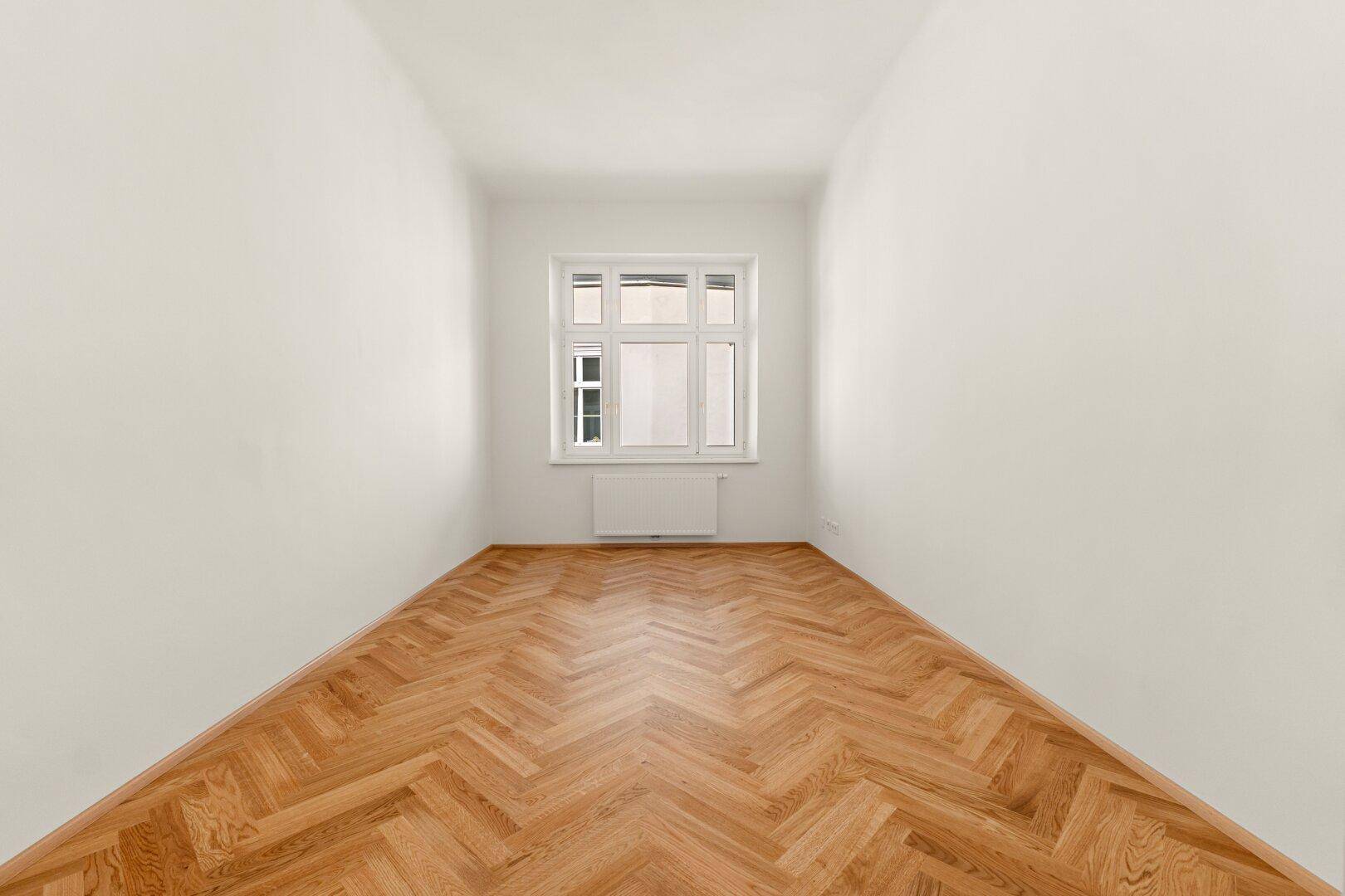 Schmaler, heller Raum mit Fischgrätparkettboden und einem Fenster, ideal als Schlaf- oder Arbeitszimmer.