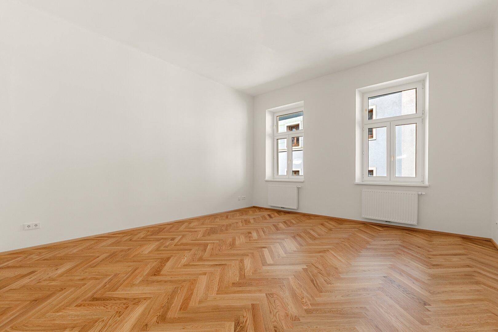 Geräumiges, helles Zimmer mit Fischgrätparkettboden und zwei Fenstern, bereit zur individuellen Gestaltung.