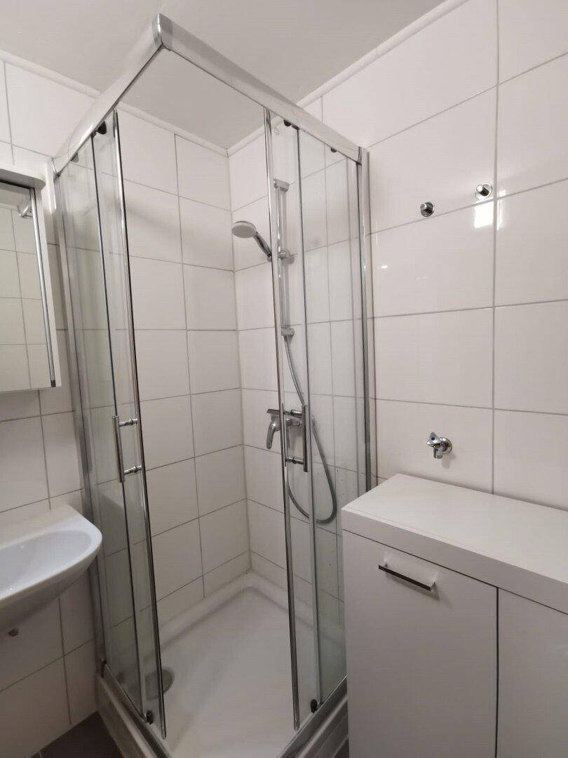 Helles Badezimmer mit weiß gefliesten Wänden, einer Eckduschkabine und einem Waschbecken mit Unterschrank.
