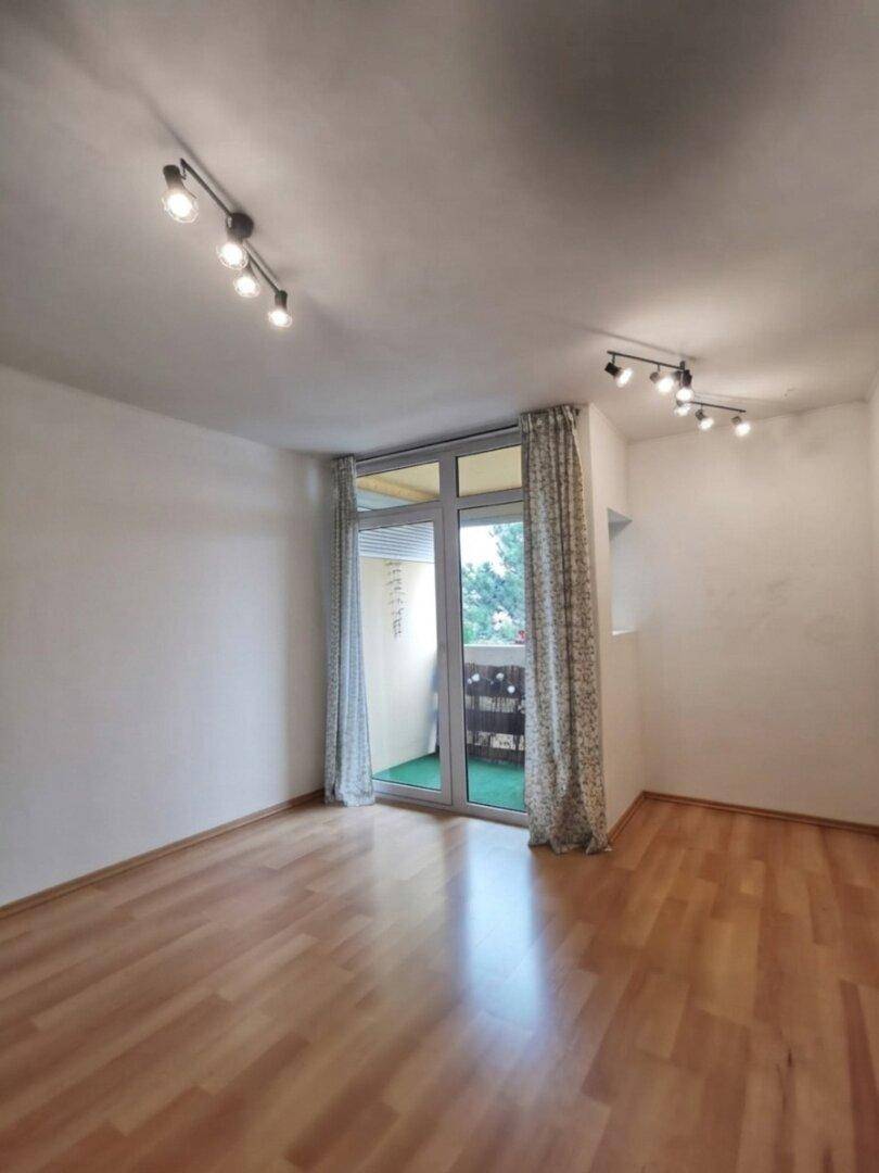Heller, leerer Raum mit Holzfußboden, weißen Wänden und Zugang zu einem Balkon.