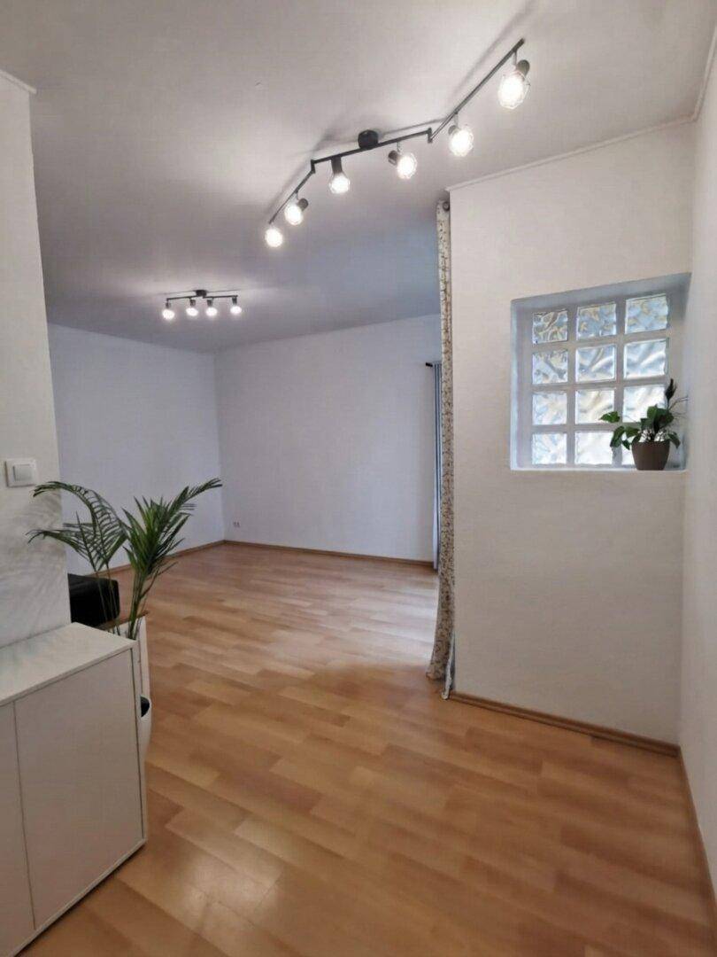 Geräumiger Wohnbereich mit hellem Holzfußboden, weißen Wänden und einem dekorativen Glasbausteinfenster.