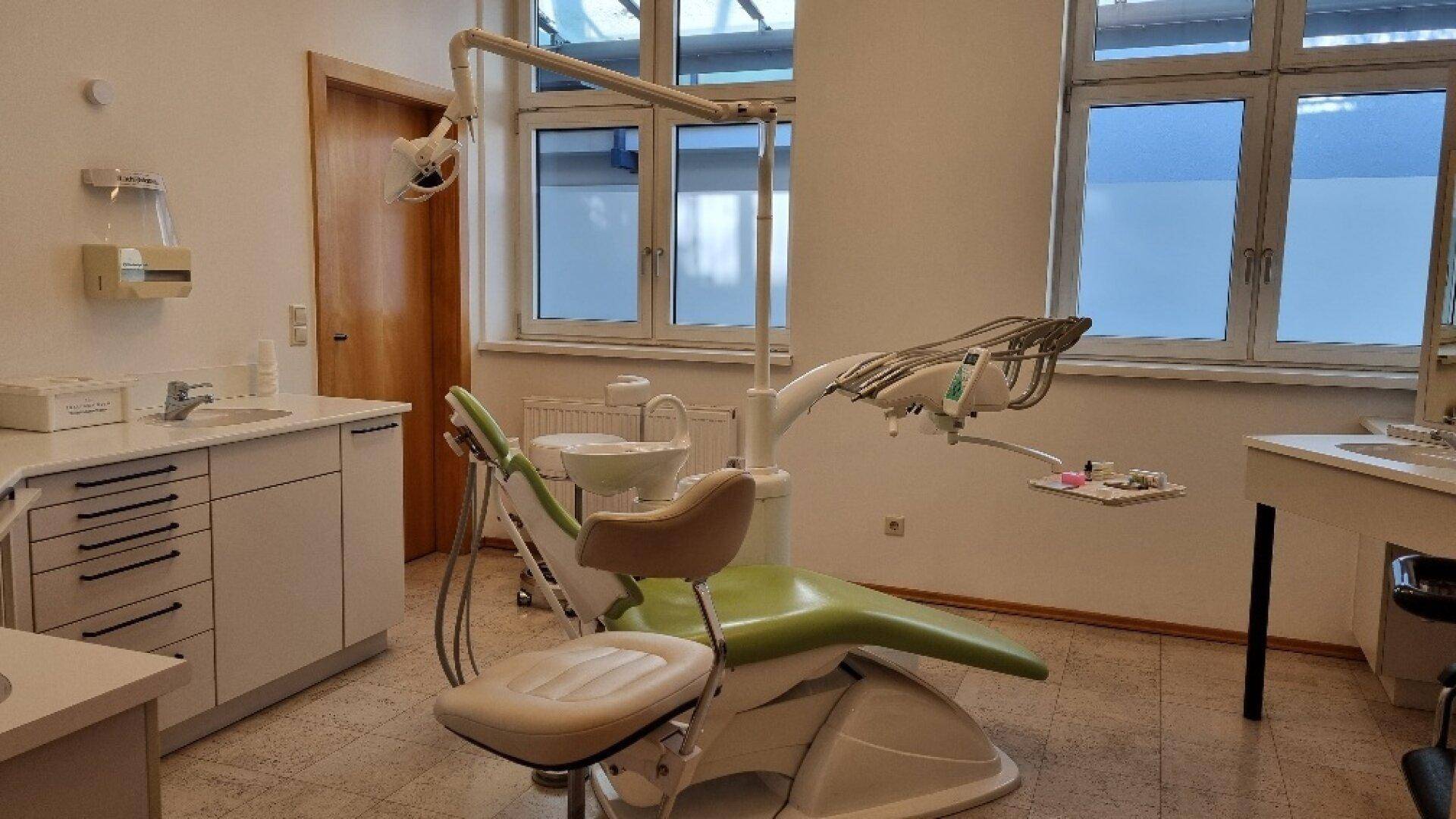 Behandlungszimmer mit zahnmedizinischer Ausstattung, weißen Schränken und großen Fenstern, hell und sauber.
